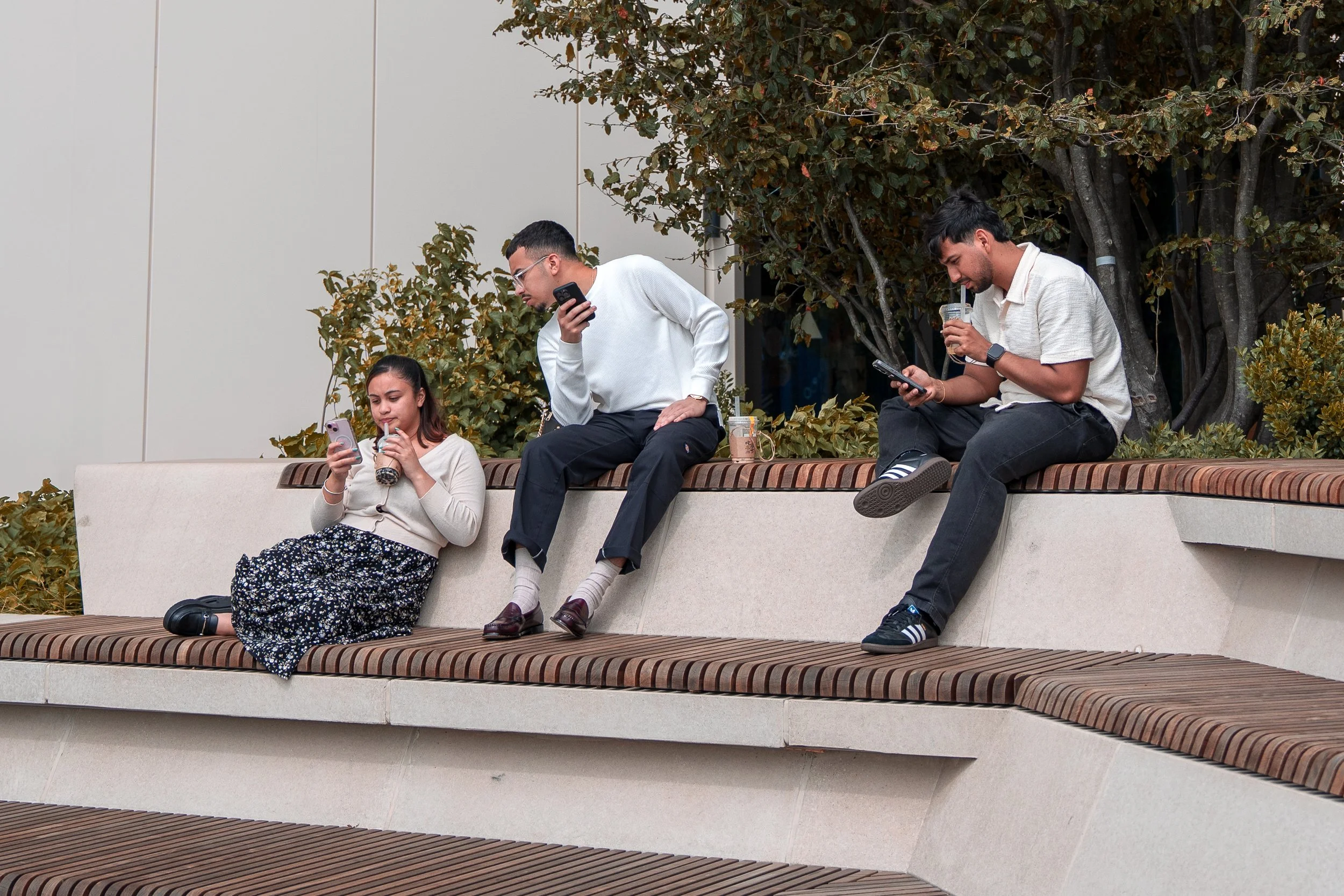 Four young people sitting on an outdoor bench, all looking at their phones and holding drinks.