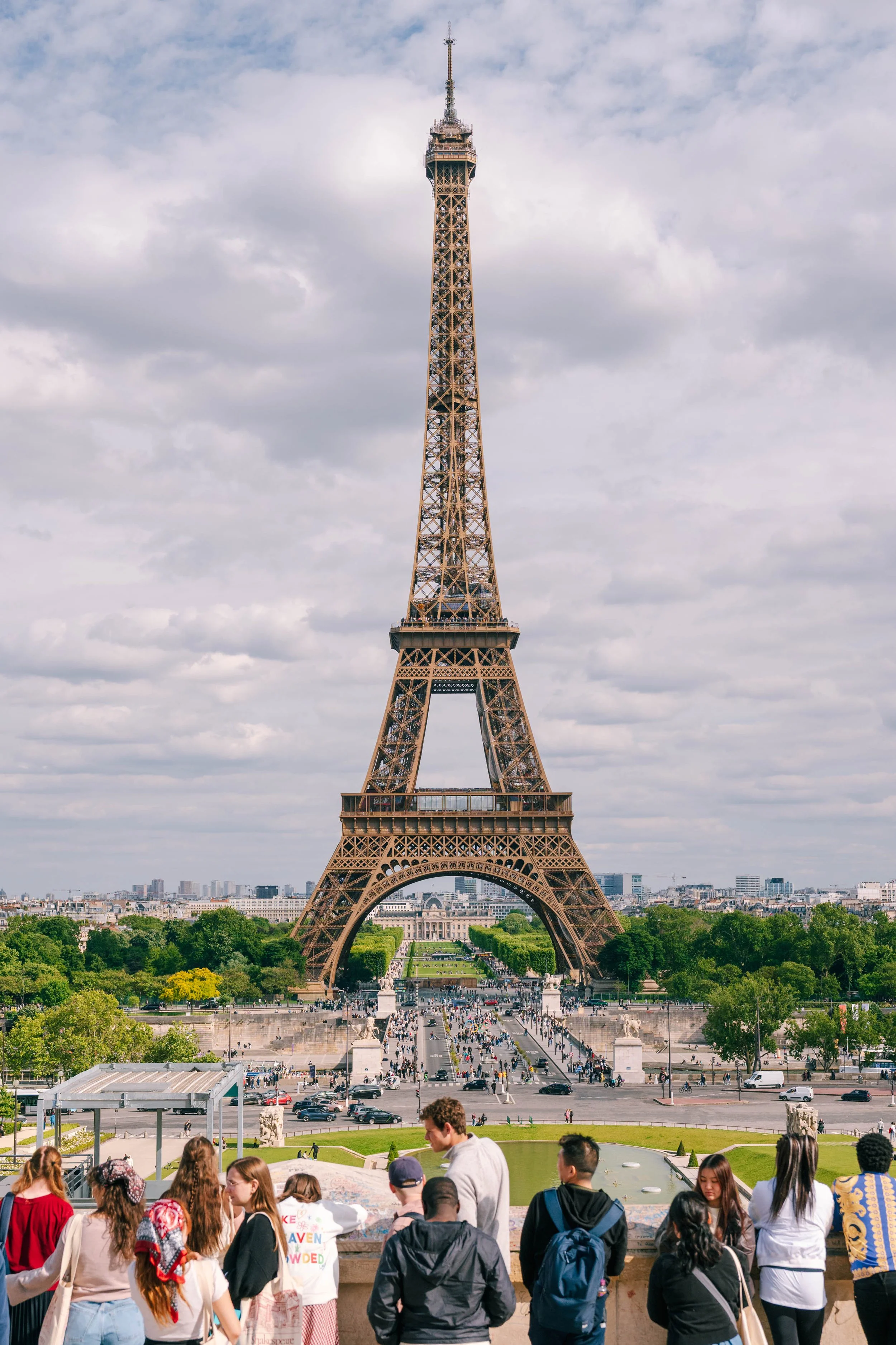 The Eiffel Tower in Paris, France, with tourists and visitors at the base, some waiting and others walking, under a partly cloudy sky.
