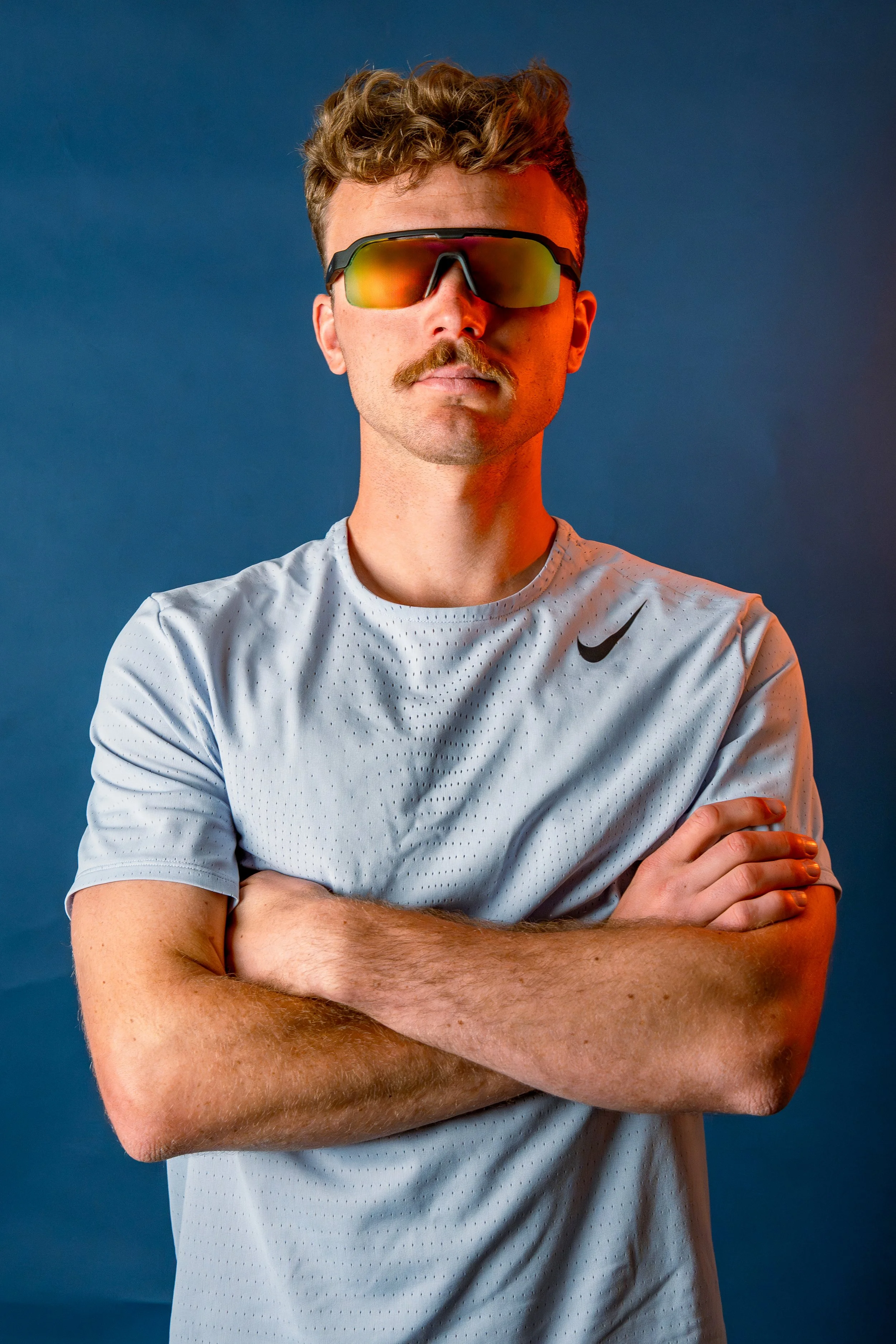 A man with a mustache and curly hair wearing reflective sunglasses and a light gray Nike athletic shirt, standing with arms crossed against a blue background.