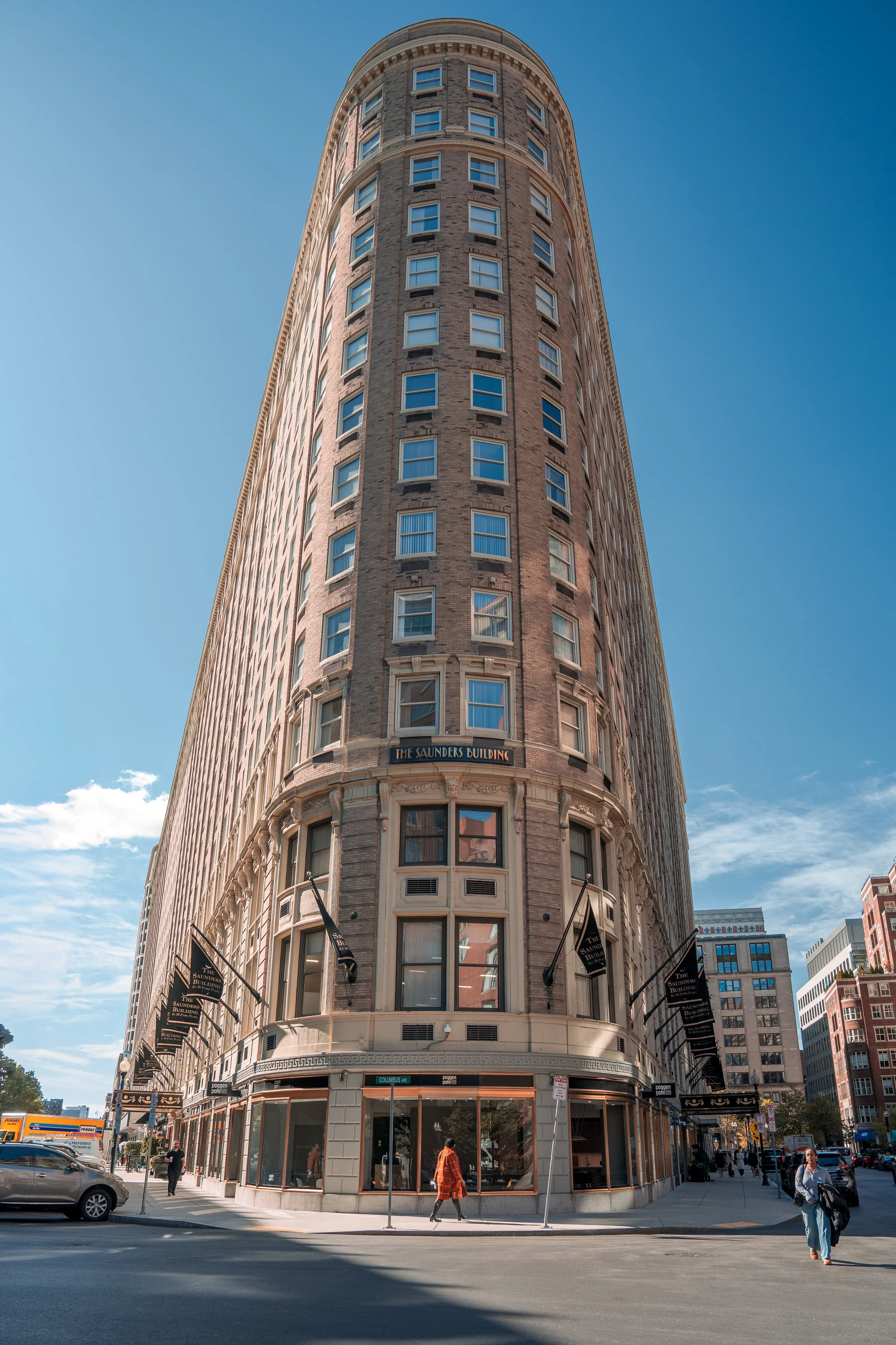 The Saunders Building, a tall, rounded corner skyscraper with brick and stone facade, black flags hanging from the exterior, and busy street scene at its base in an urban environment during daytime.