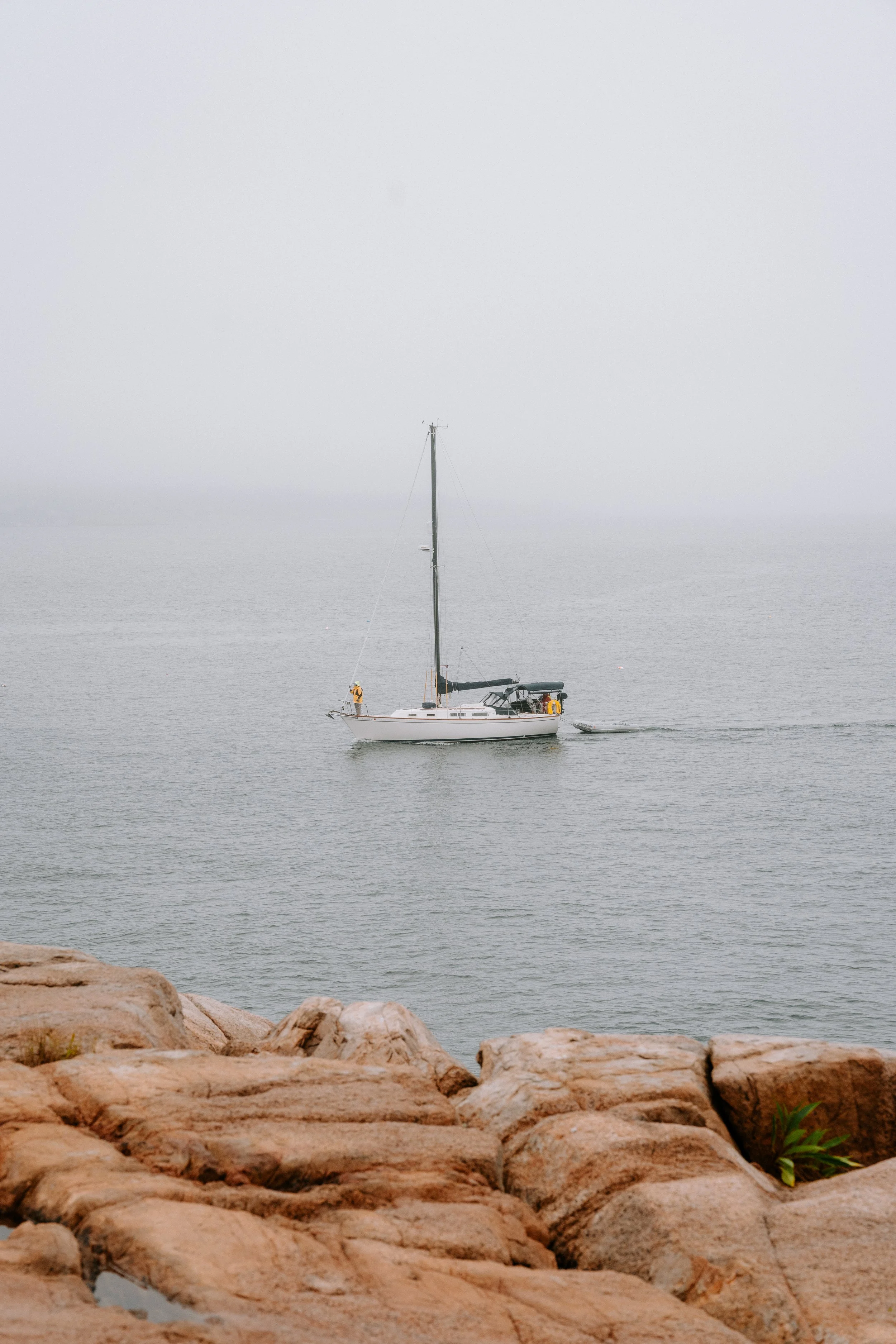 A sailboat floating on calm water viewed from a rocky shoreline with foggy background.