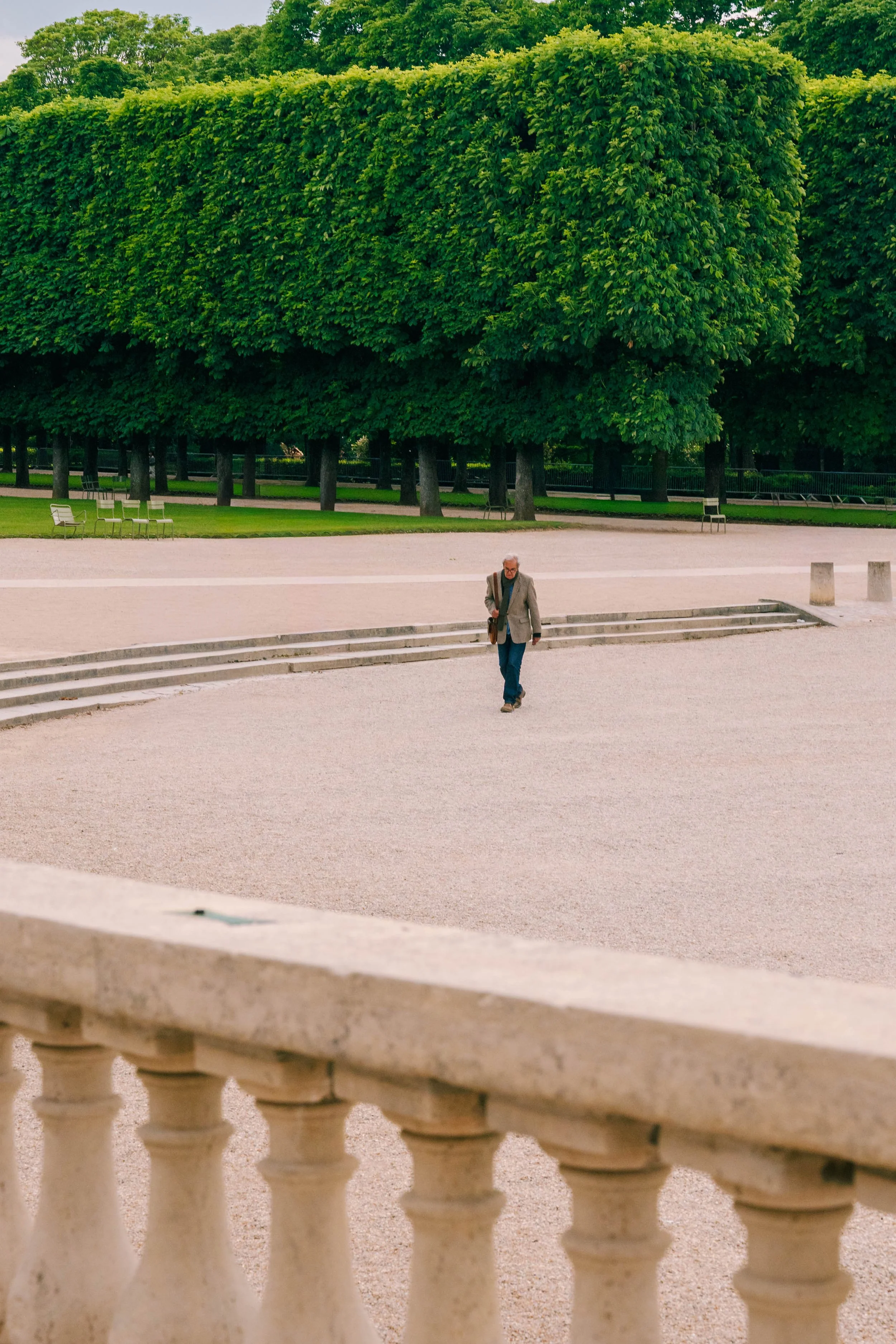 A man with gray hair and glasses walks alone in a park with lush green trees in the background. The scene is viewed from behind a stone balustrade.