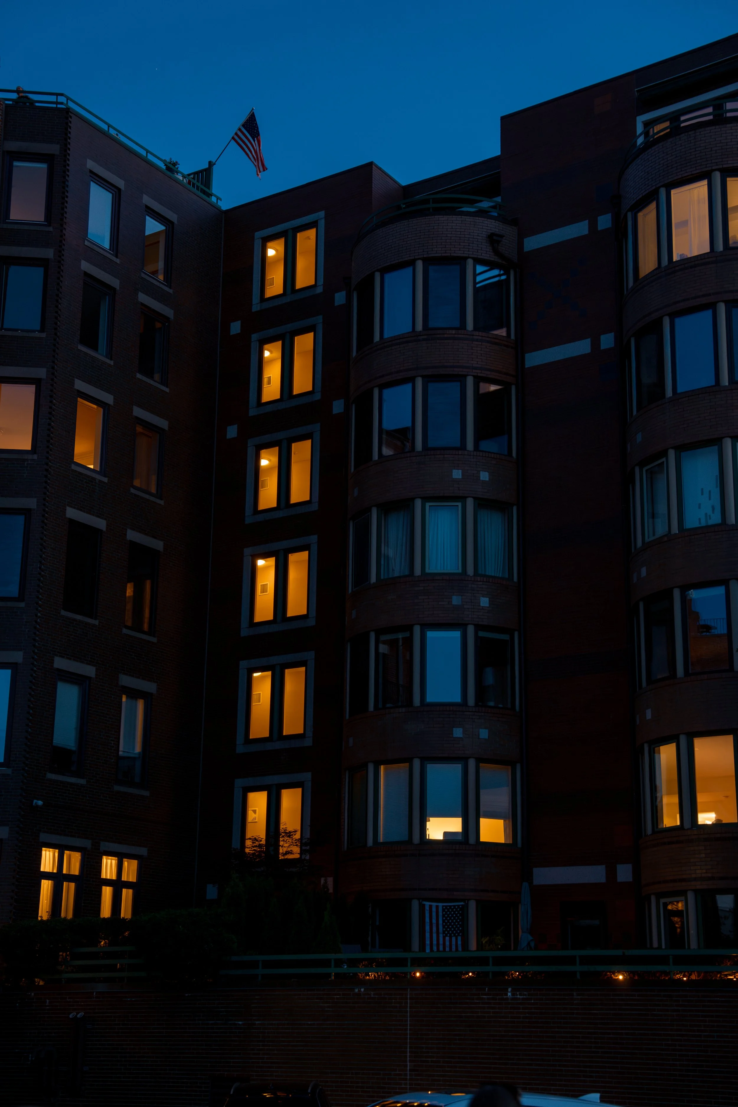 Apartment building at dusk with some windows lit and a small American flag on the roof.