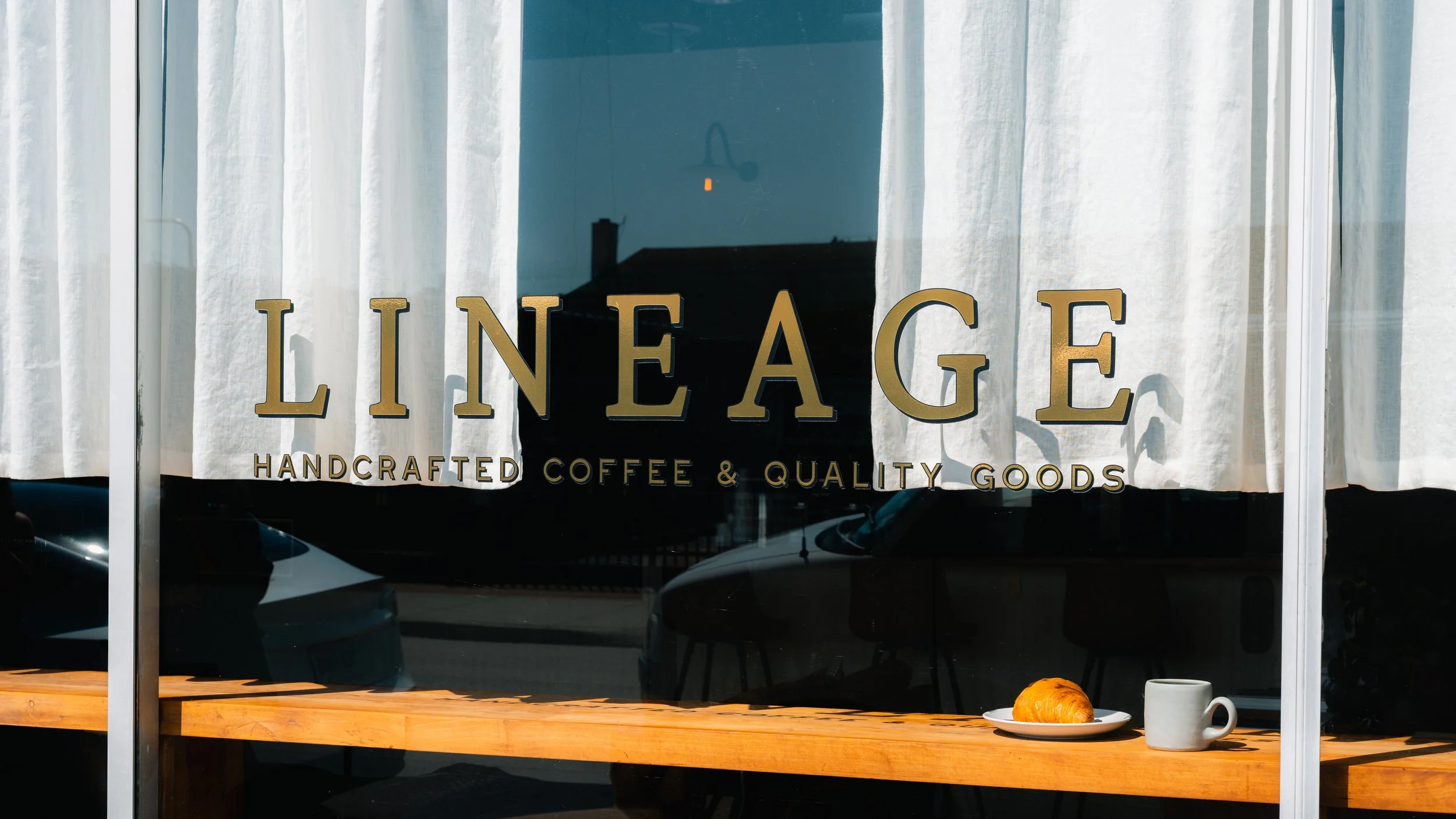 A storefront window with white curtains, a sign reading 'LINEAGE Handcrafted Coffee & Quality Goods,' and a wooden table displaying a plate with a croissant and a ceramic mug.