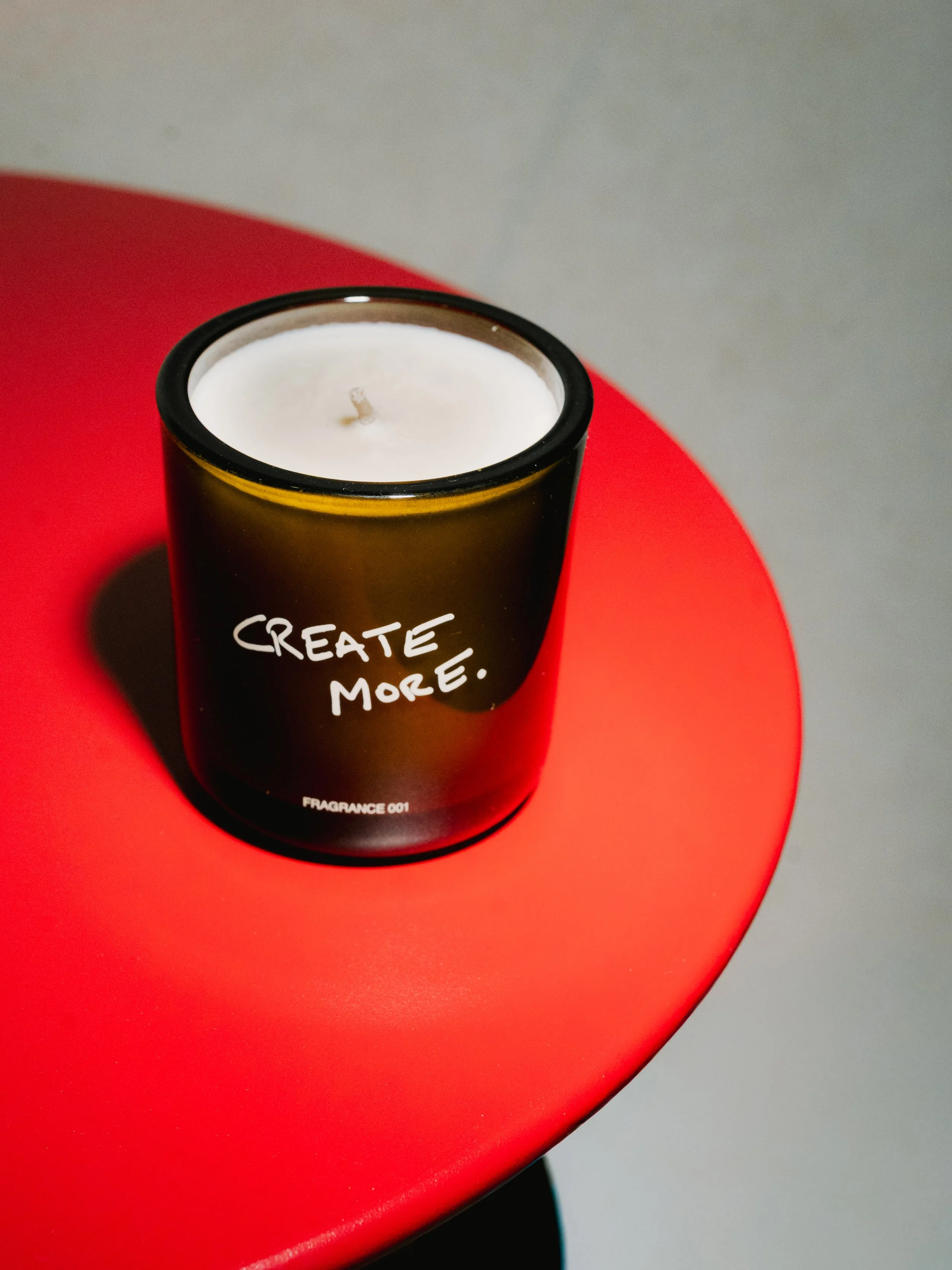 A black candle jar with white wax and a wick, placed on a red circular table, with the words 'CREATE MORE.' written on it.