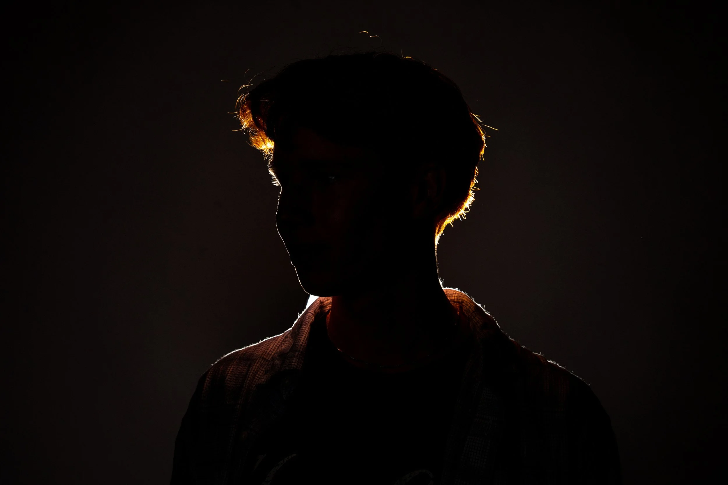 Silhouette of a person with backlit hair and face in shadow, wearing a checkered shirt, against a dark background.
