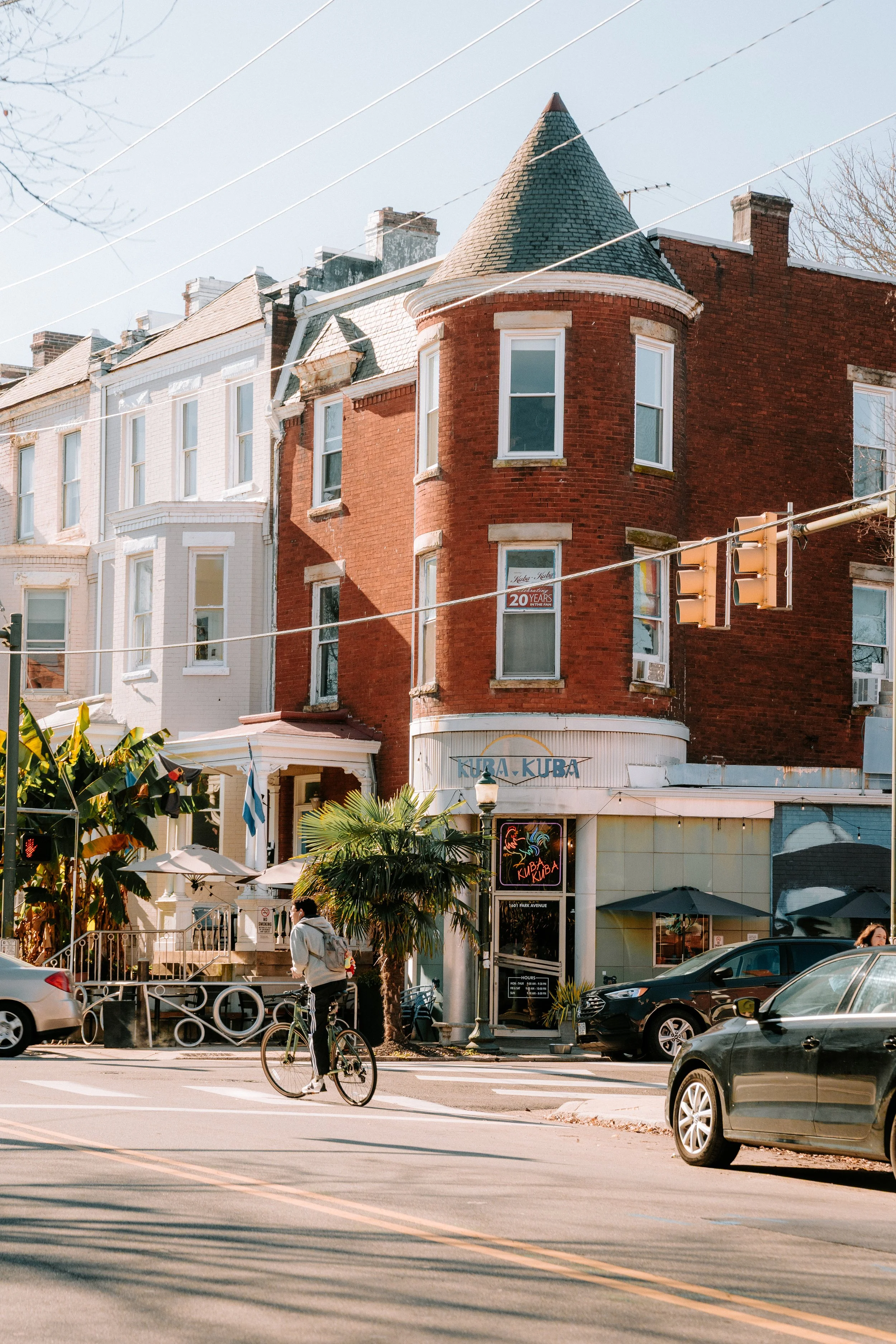 A street scene with a red brick building featuring a rounded turret with a conical roof. The ground level has a restaurant called Kubar Kubar with neon signs. A person is riding a bicycle in the foreground, and cars are parked along the street. There are some palm trees and umbrellas outside the restaurant, and overhead power lines are visible.