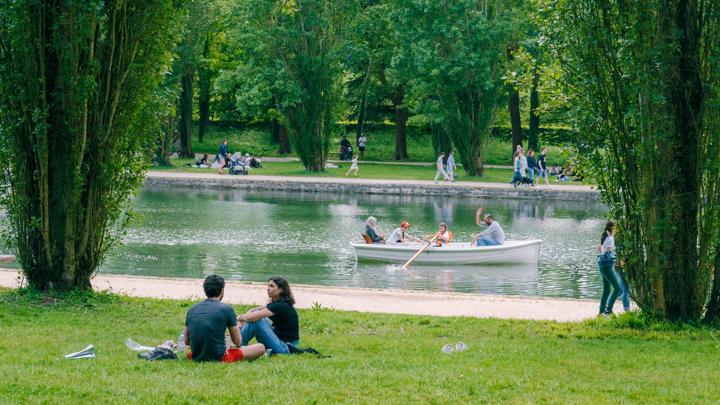 People enjoying a day at the park by a pond, with some rowing a boat and others relaxing on the grass and walking along paths, surrounded by green trees.
