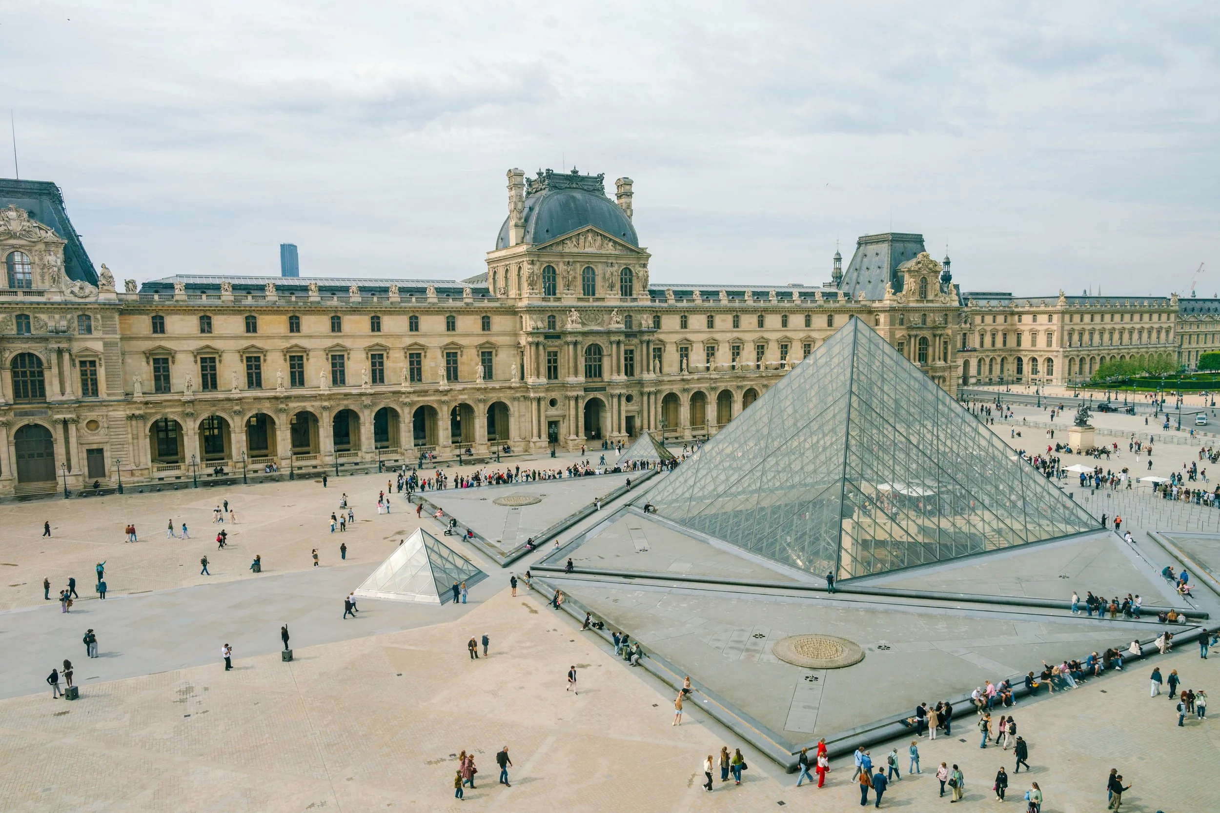 View of the Louvre Museum in Paris, France, with glass pyramids in the foreground and historical buildings in the background, filled with visitors.