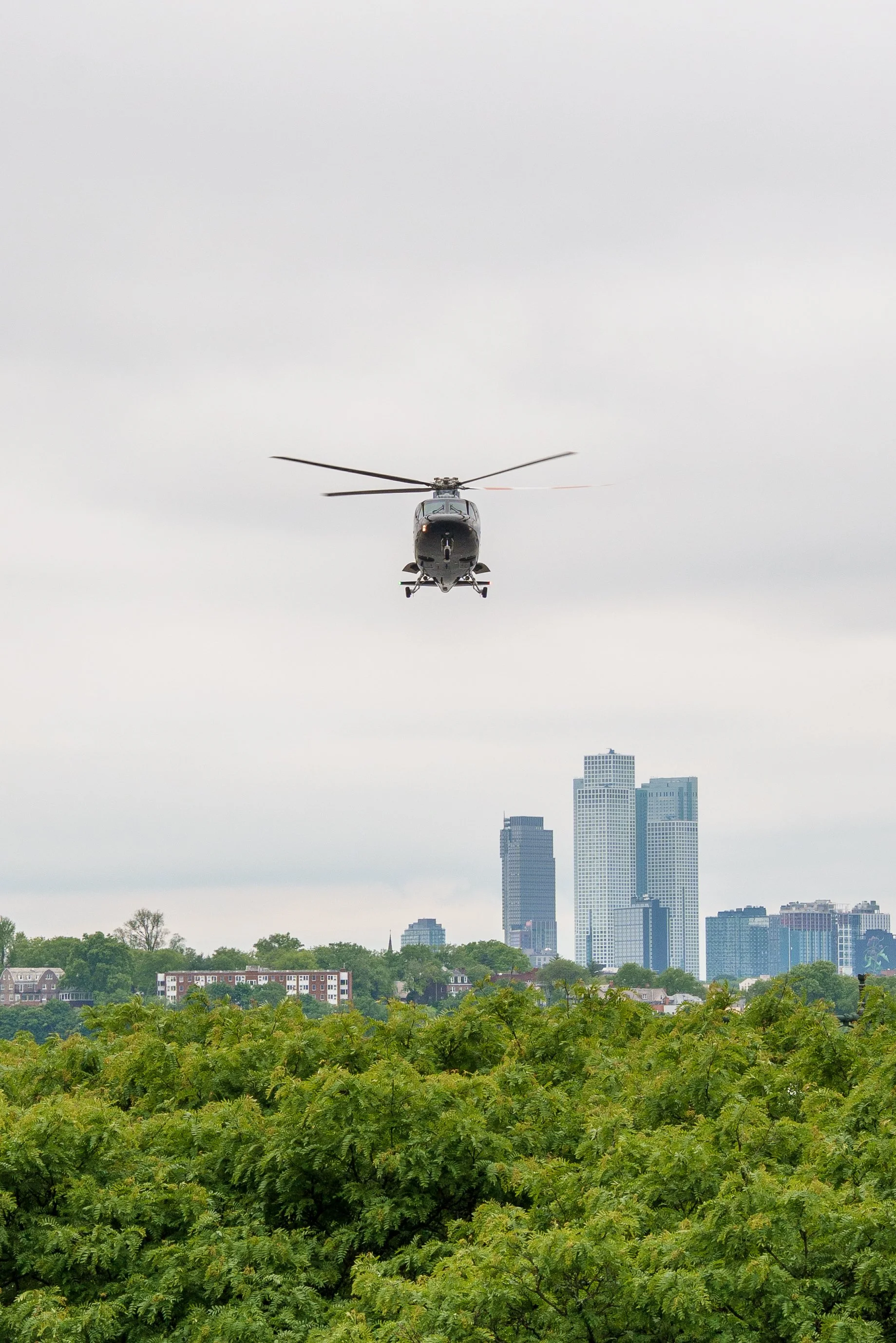 A helicopter flying over a cityscape with trees and tall buildings, under an overcast sky.
