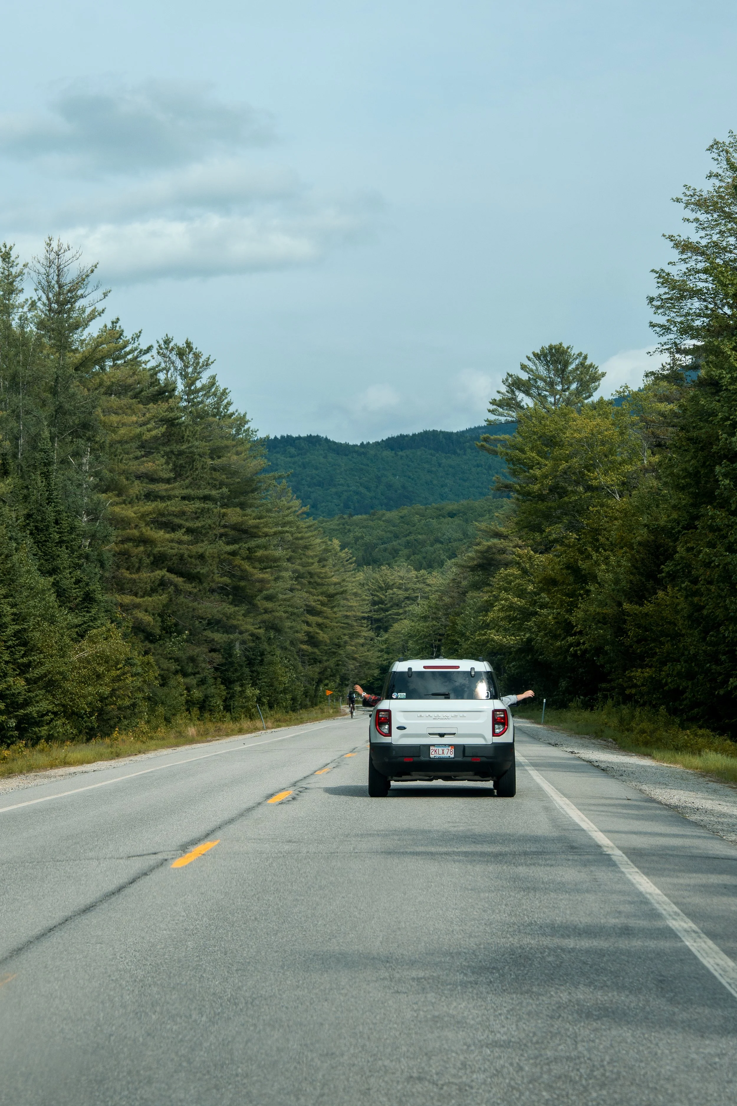 A vehicle on a two-lane rural road surrounded by green trees on both sides, with hills in the background and a cloudy sky overhead.