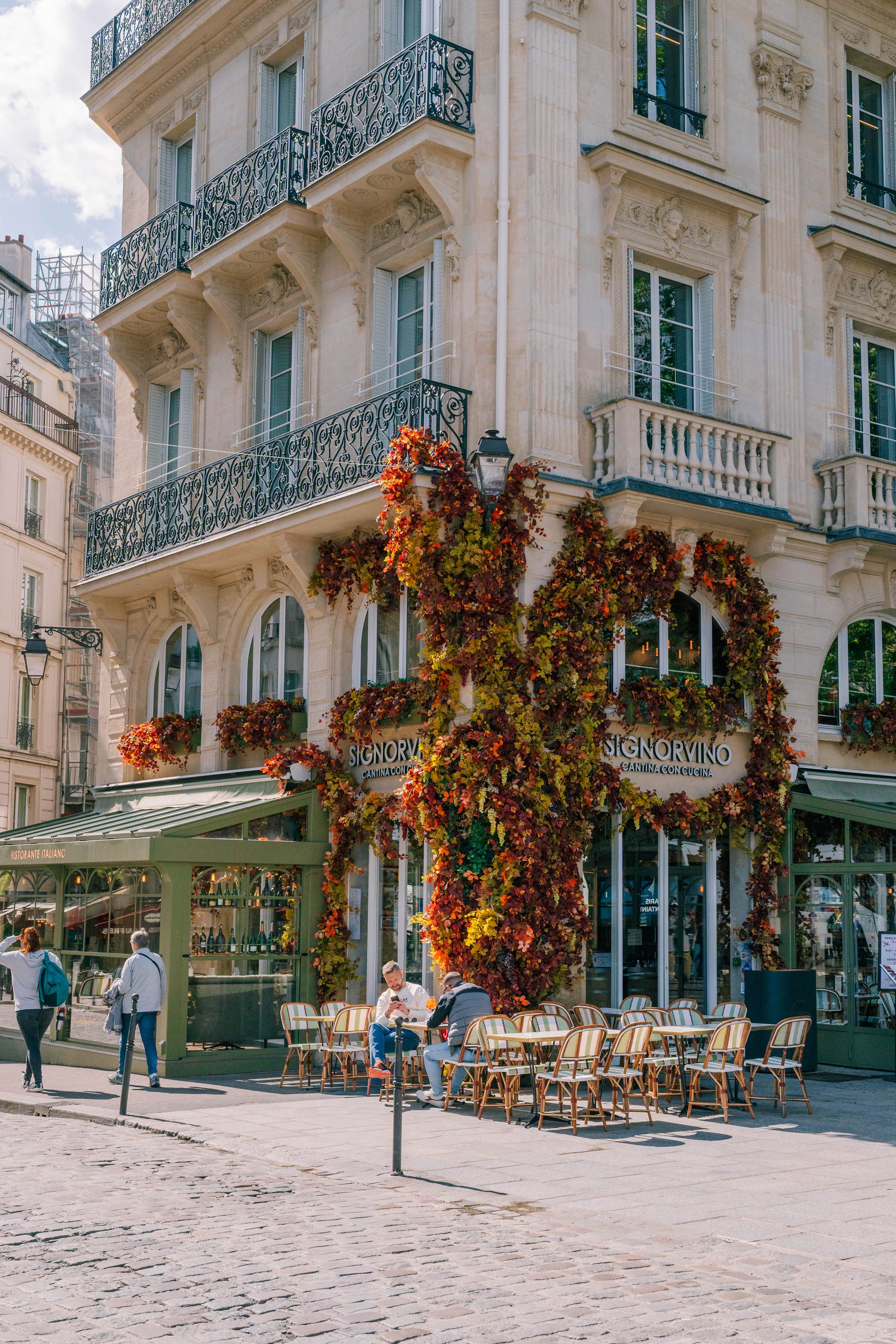 Parisian building with ornate balconies and a restaurant on the ground floor called SIGNORVINO with an outdoor seating area filled with colorful autumn leaves and people sitting and walking on the cobblestone street.