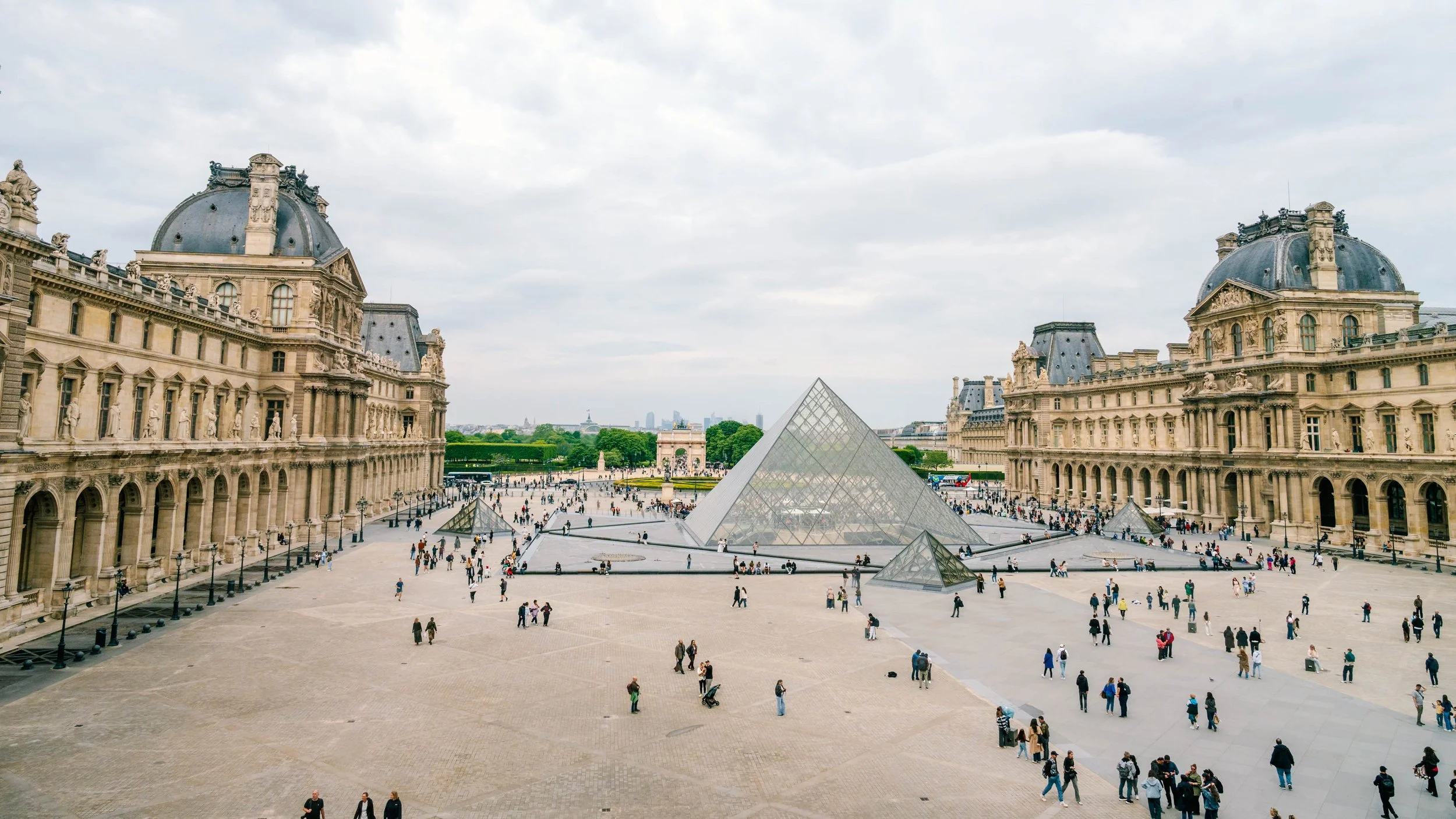 View of the Louvre Museum courtyard in Paris, France, showing glass pyramids and historic buildings with sculptures on the rooftops, with many visitors walking around.
