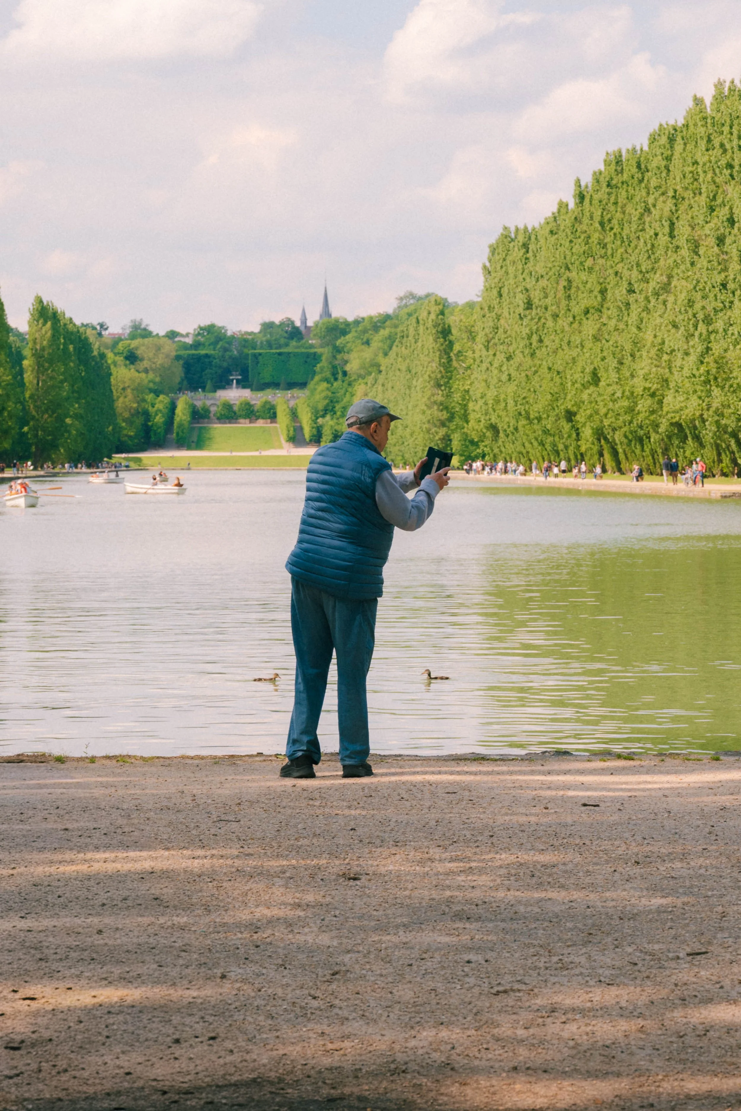An elderly man in a blue vest and gray cap stands by a lake, looking at and possibly reading from a small book or device, with trees and a park in the background and people walking along the pathway.