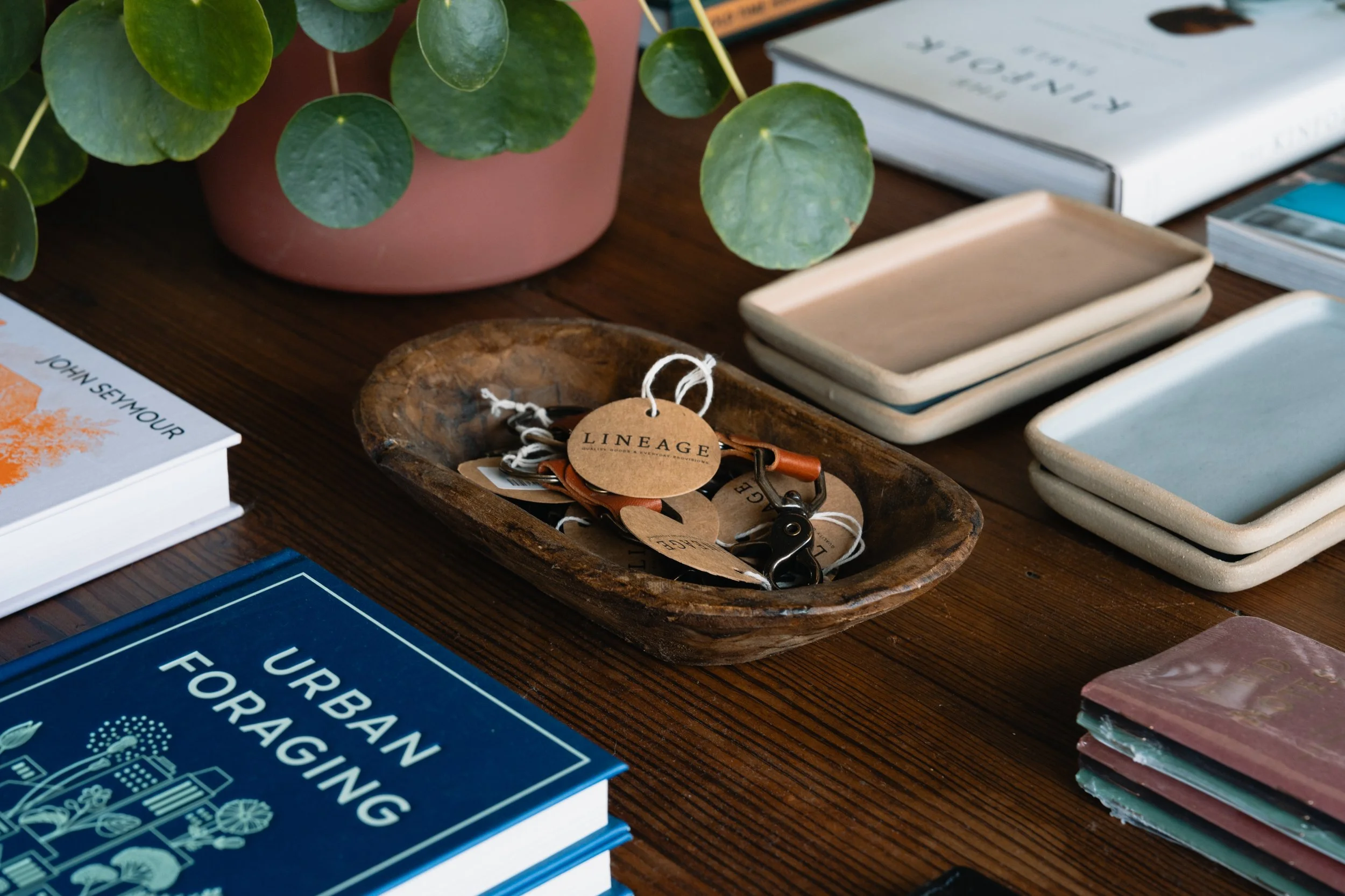 Close-up of a wooden table with a potted plant, a wooden bowl with keychains, a stack of ceramic trays, and books about urban farming and aquaponics.