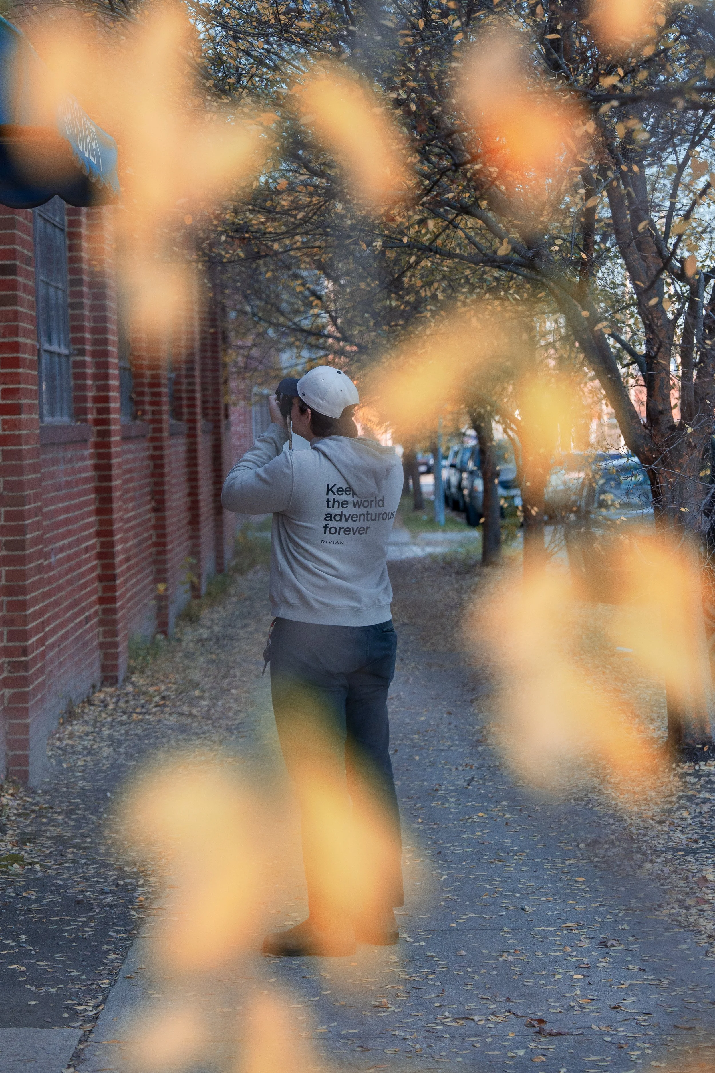 A person wearing a gray hoodie and a baseball cap, holding binoculars, stands on a sidewalk amid autumn leaves and trees, with a red brick building and parked cars in the background.