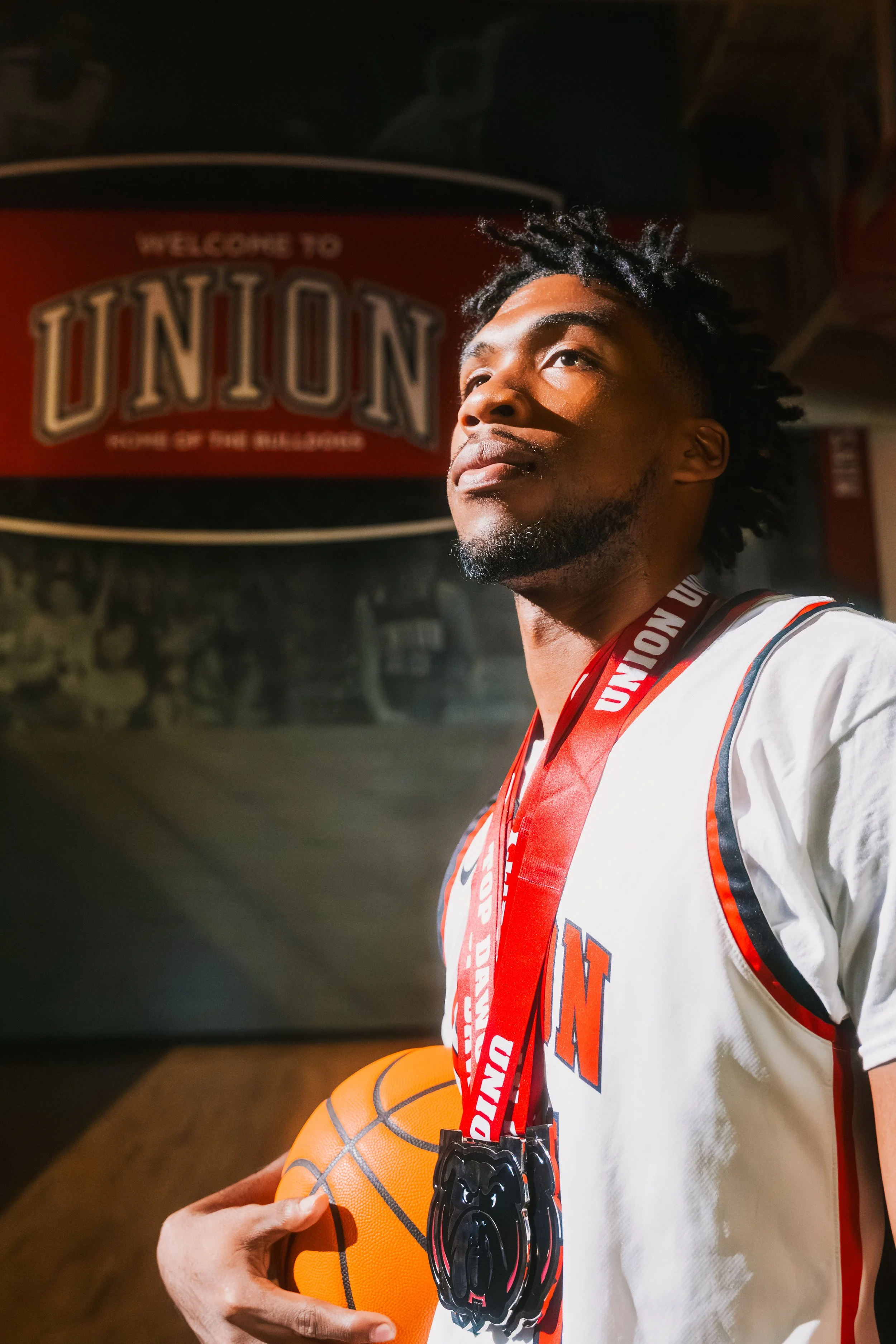 Young man with dreadlocks holding a basketball and wearing a medal around his neck, standing in front of a sign that says 'Welcome to Union'.