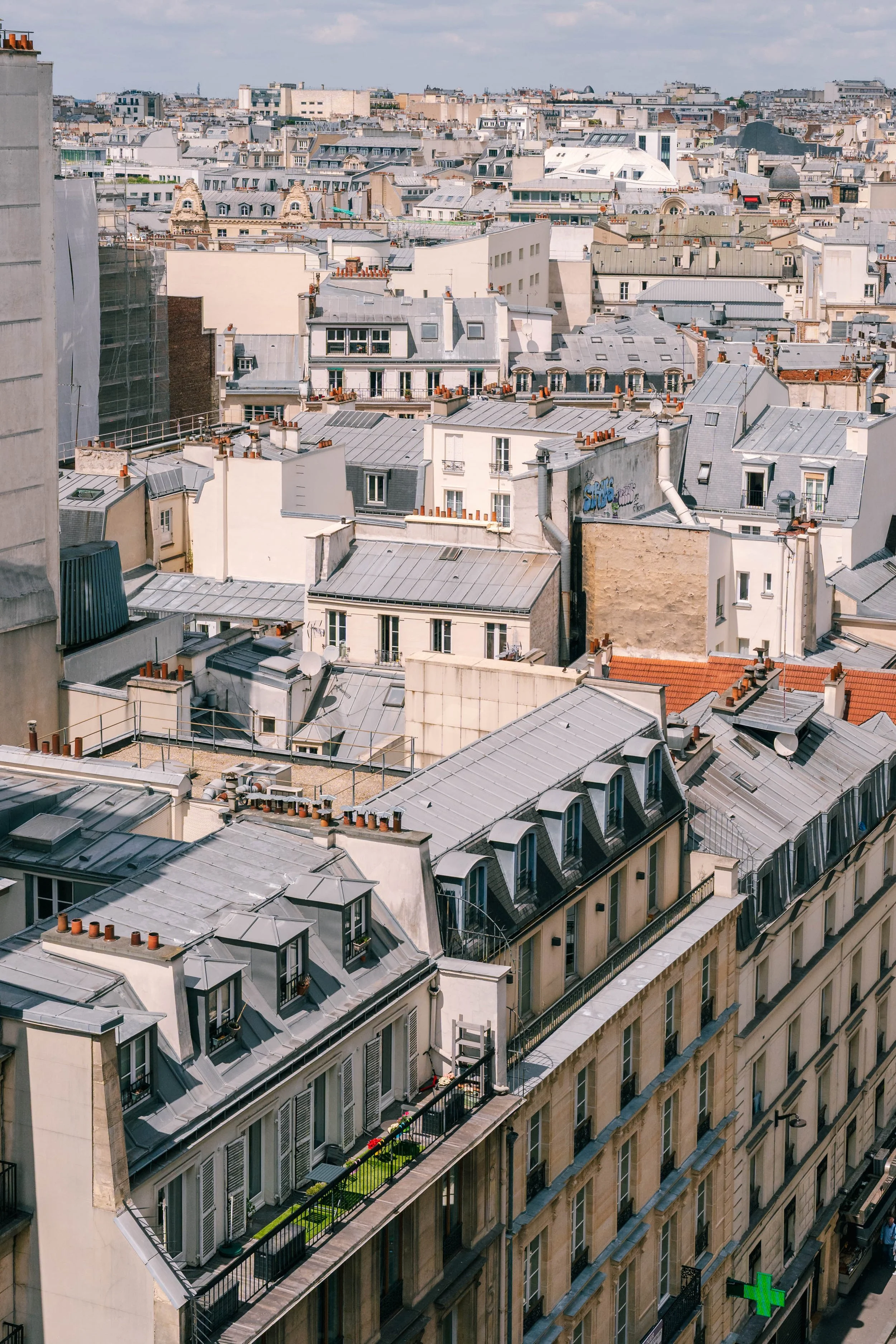 Paris city rooftops with mansard roofs, chimneys, and laundry hanging on a balcony, with a pharmacy green cross sign on the street below.