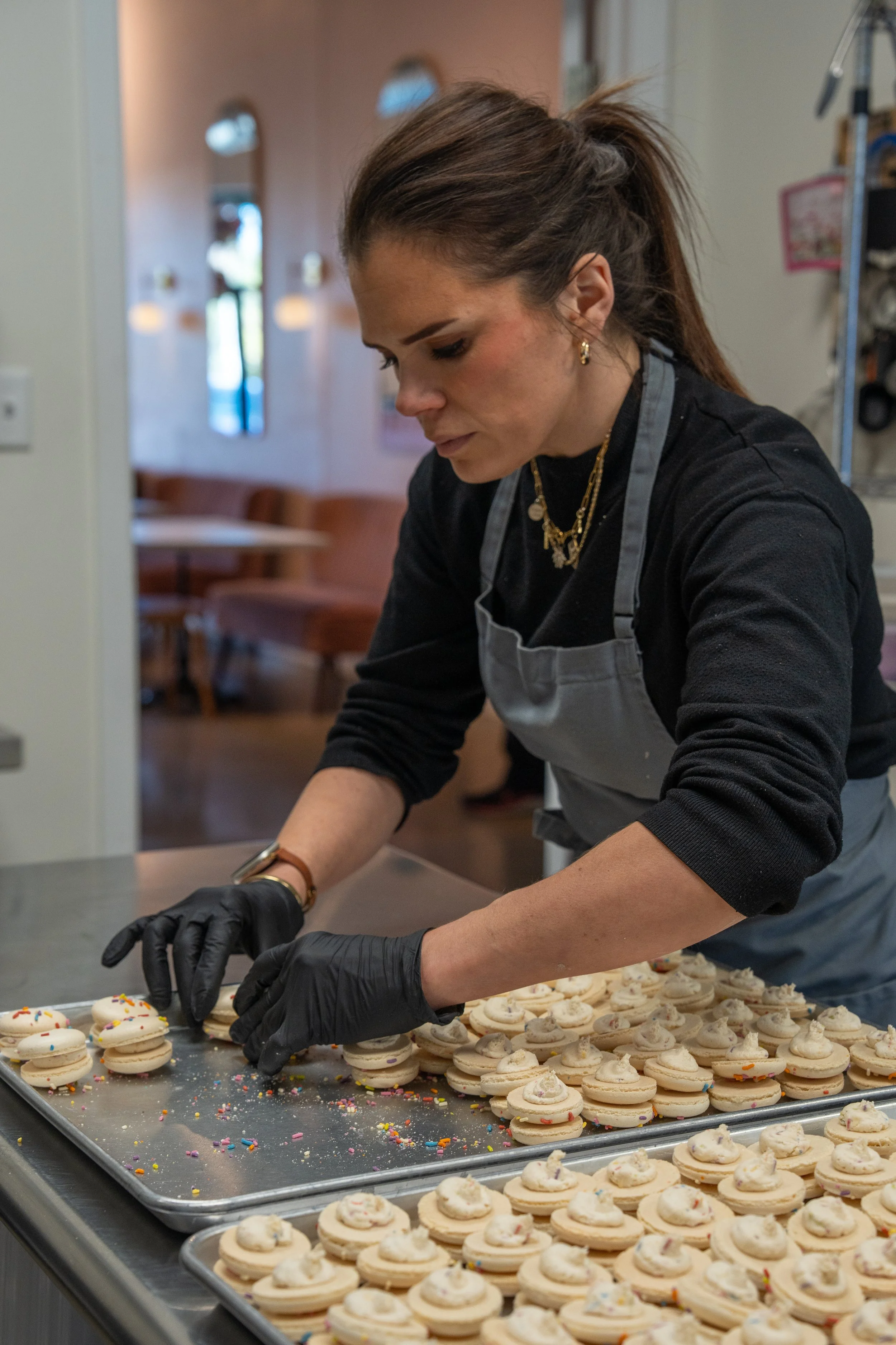 A woman in a black shirt and gray apron, wearing black gloves and gold jewelry, is preparing cupcakes with white frosting and colorful sprinkles on a large tray in a kitchen or bakery setting.