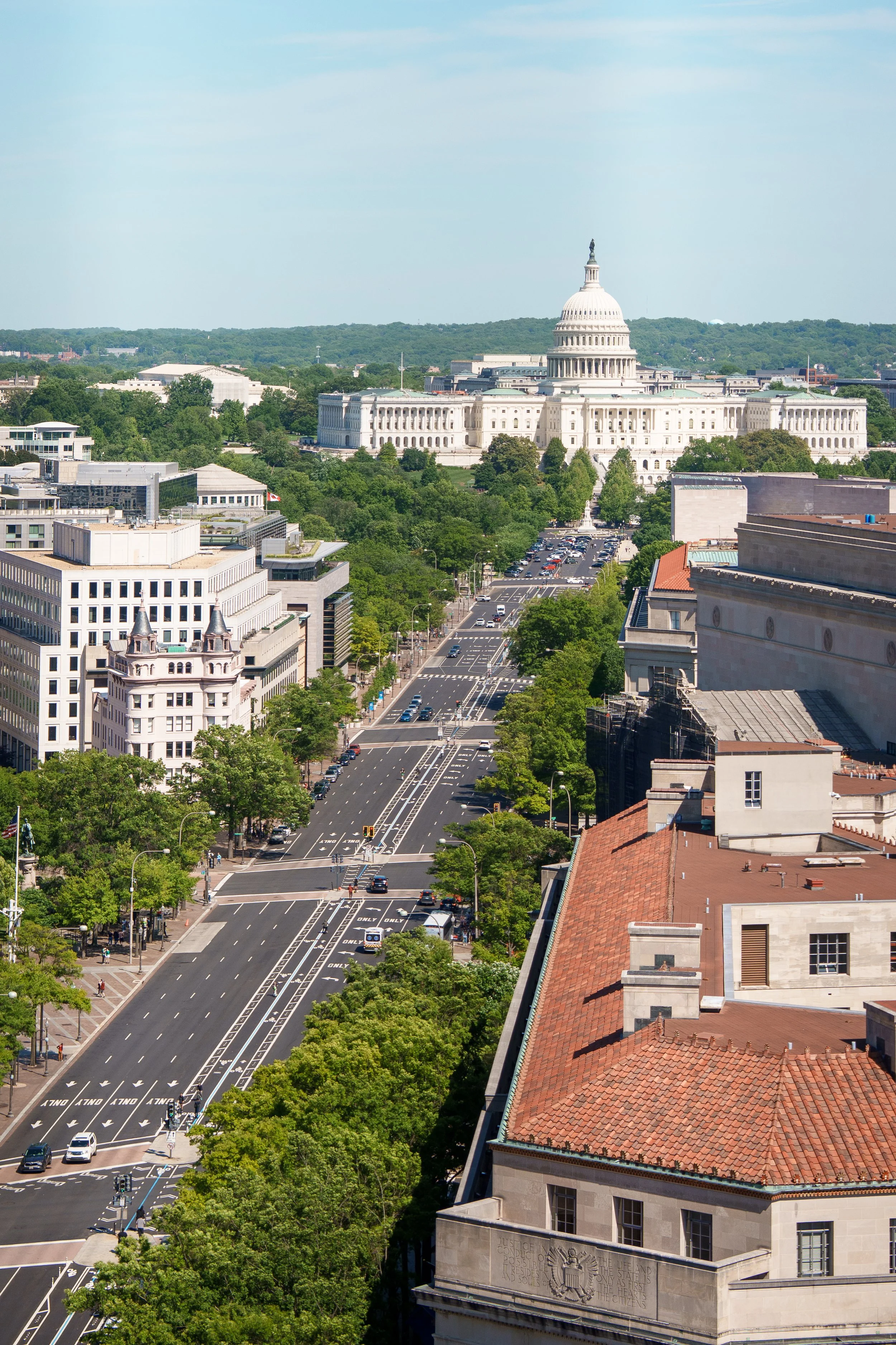 City street leading towards the U.S. Capitol building in Washington D.C., with trees lining the road and buildings on both sides, under a clear blue sky.