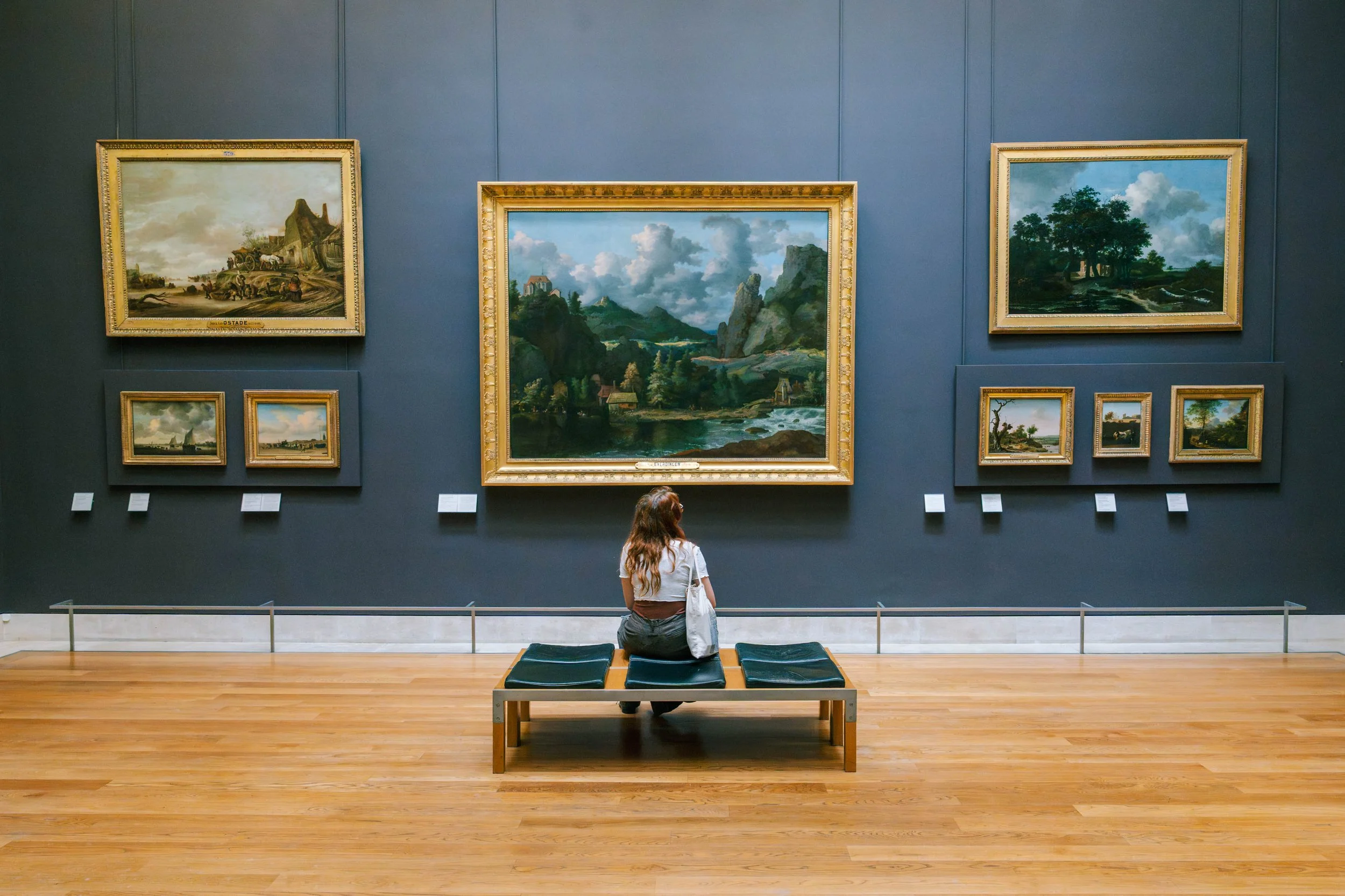 A woman sitting on a bench in an art gallery, viewing framed landscape paintings on a blue wall.