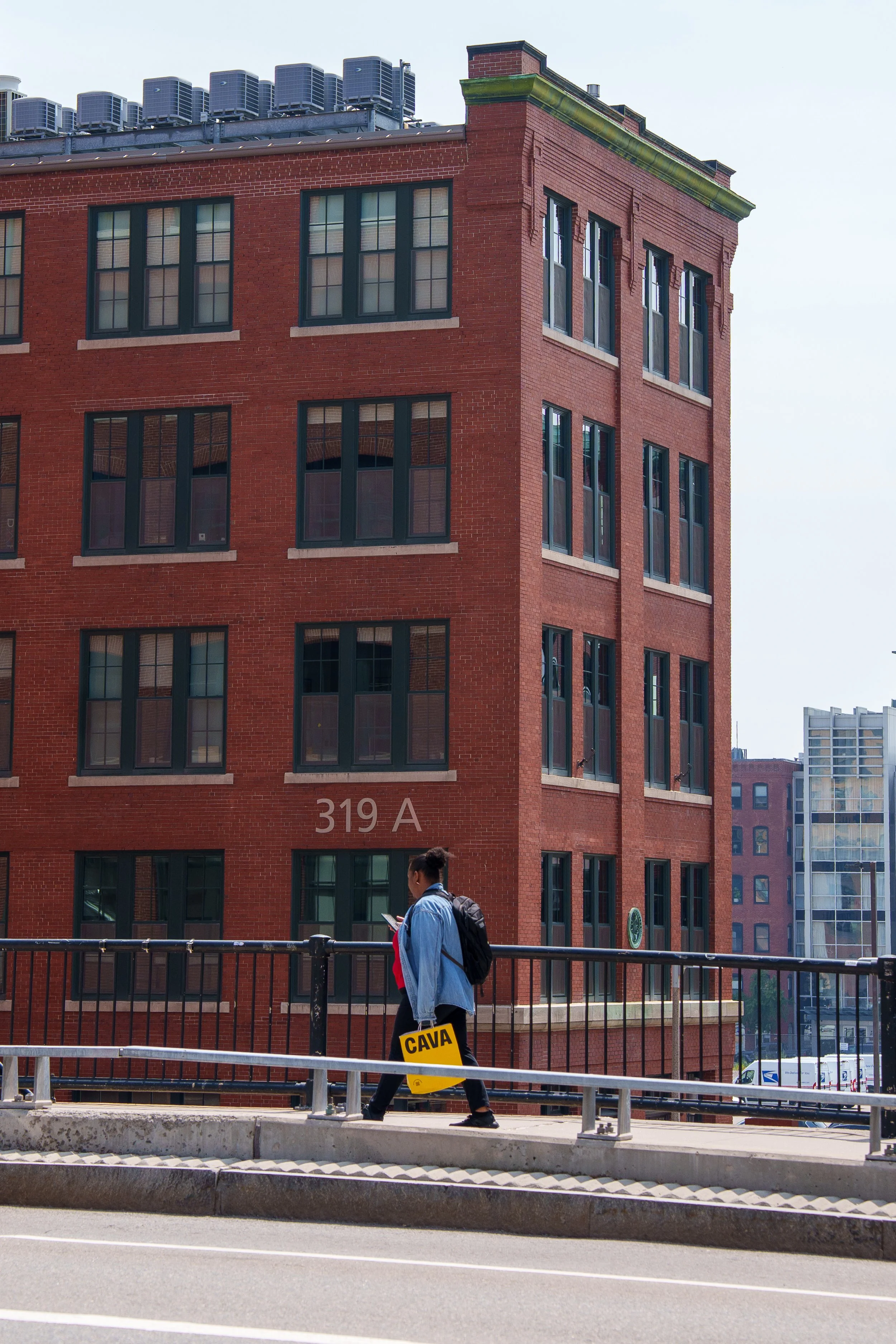 Person walking along a city street, carrying a yellow bag with the word 'CAVA' on it, with a red brick building in the background labeled '319 A'.
