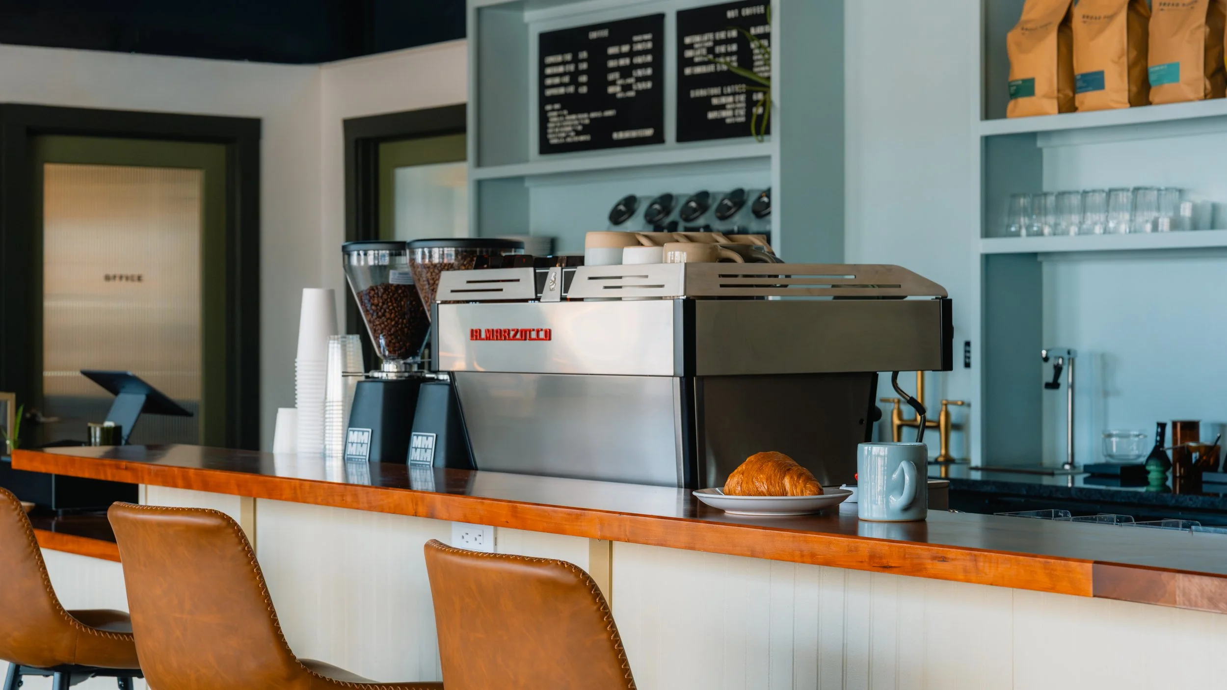Coffee shop counter with an espresso machine, a croissant on a plate, a mug, and seating with leather chairs in the foreground.