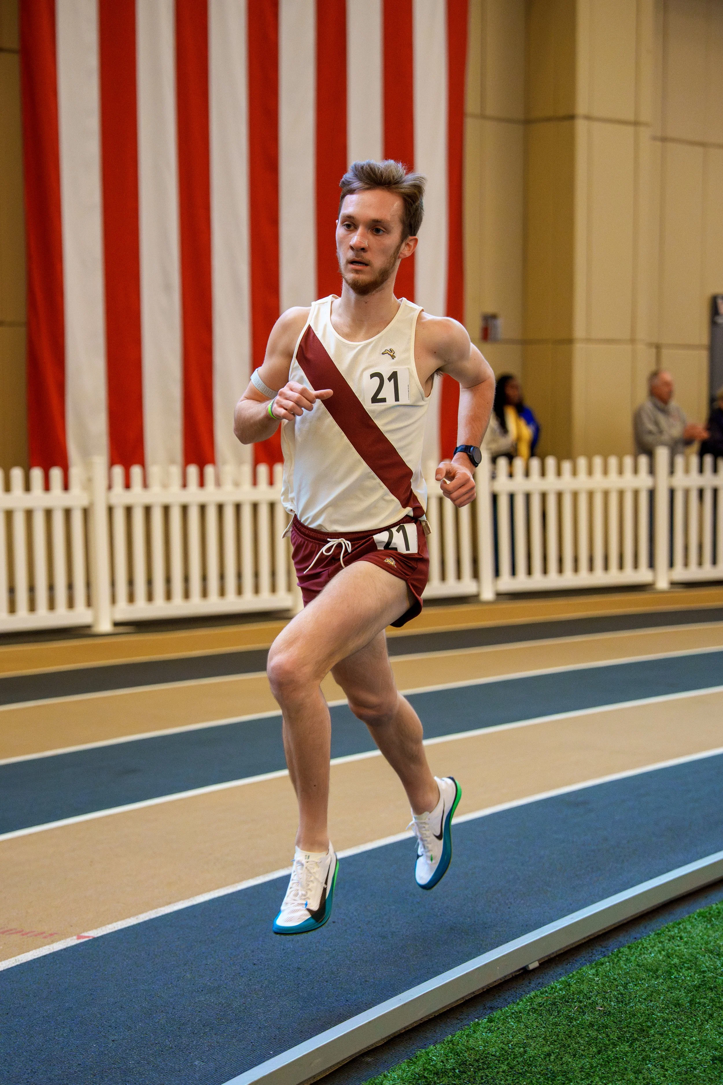 A male runner wearing a cream and maroon sports uniform with the number 21 on his chest is running indoors on a track. Behind him is a patriotic American flag and a white picket fence, with some spectators standing in the background.