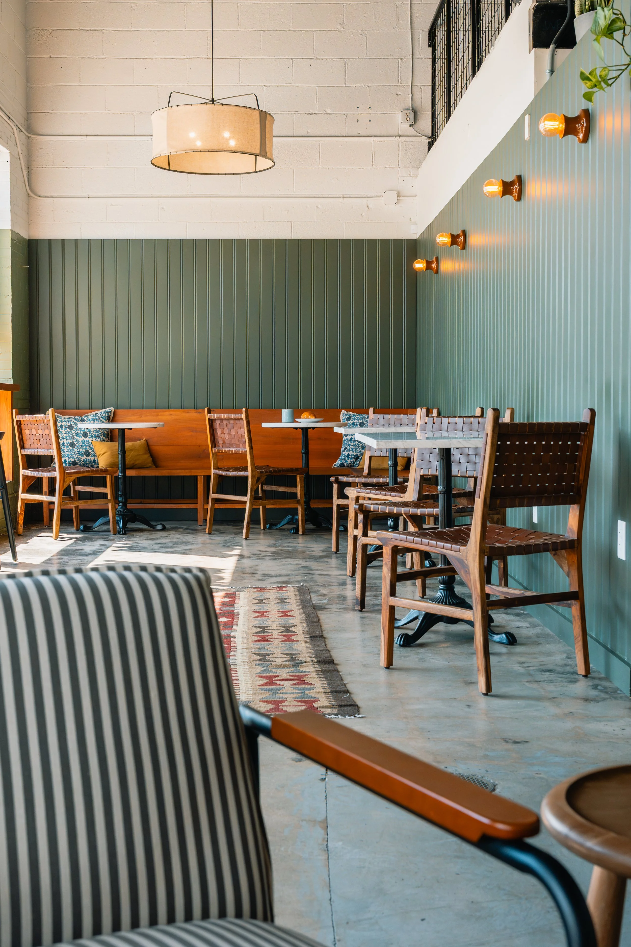 Interior of a cafe or restaurant with wooden chairs, a bench with cushions, marble tables, wall-mounted light bulbs, and a decorative rug on a concrete floor.