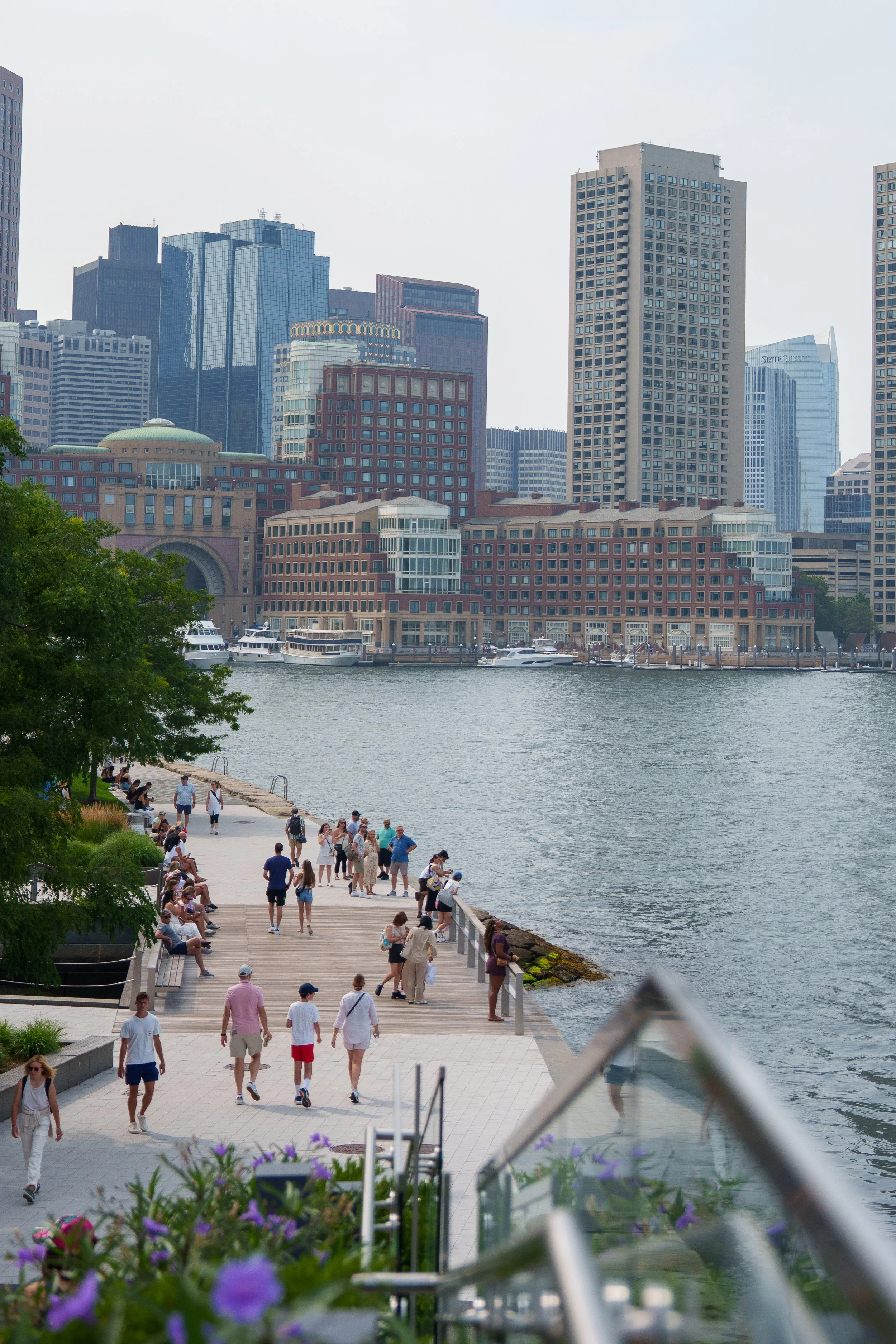 People walking and sitting on a waterfront boardwalk with high-rise buildings in the background