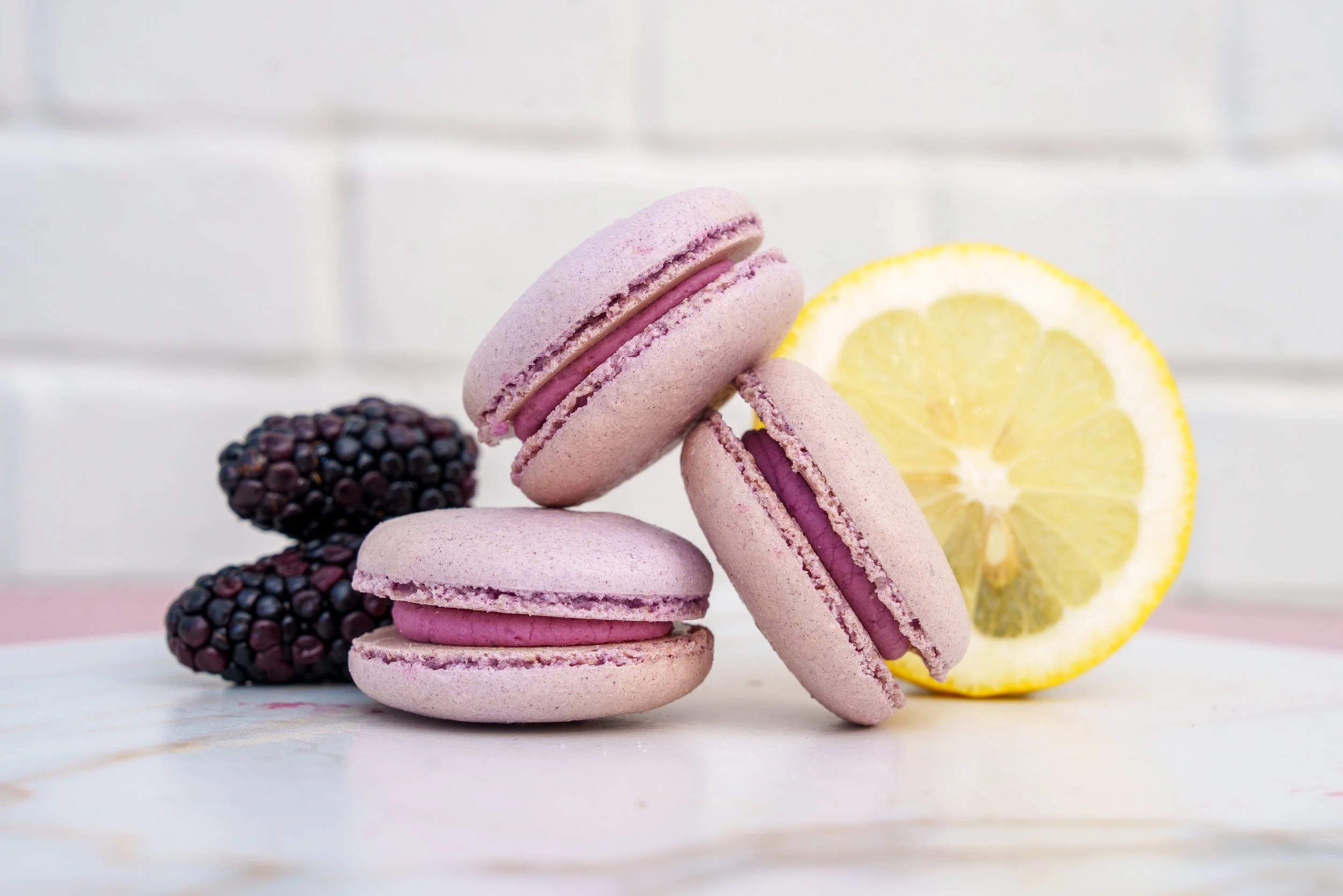A stack of pink macarons with purple filling, blackberries, and lemon slices on a white surface against a white brick wall background.