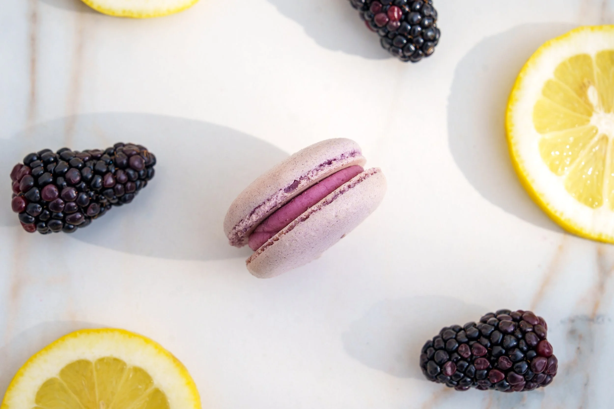 A flat lay of blackberries, lemon slices, and a pink macaron on a white surface.
