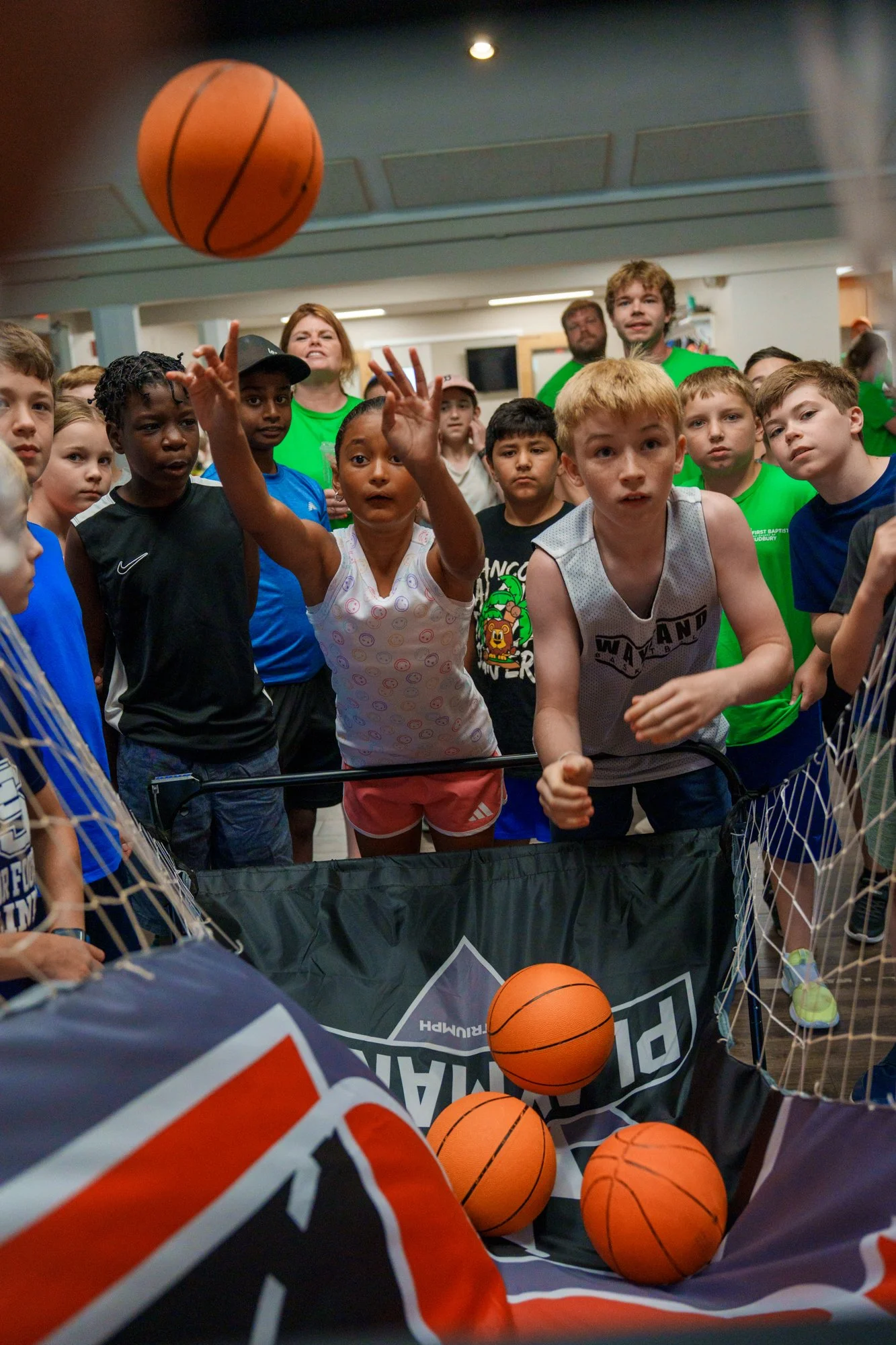 Children playing a basketball game aimed at shooting balls into a large basket at an indoor event, with a crowd watching.