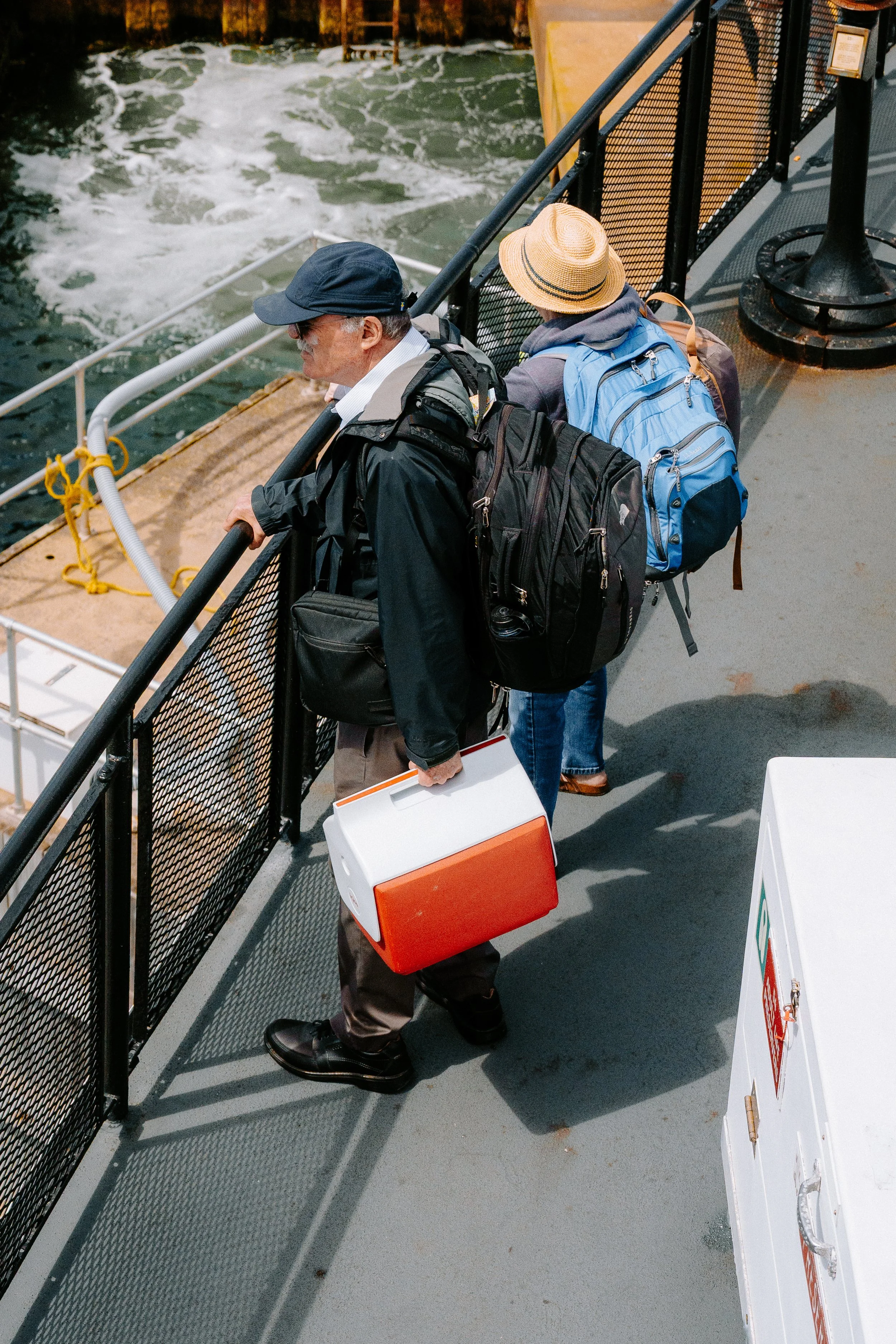 Two people with backpacks and luggage on a ferry or boat deck, looking over the water near a dock.
