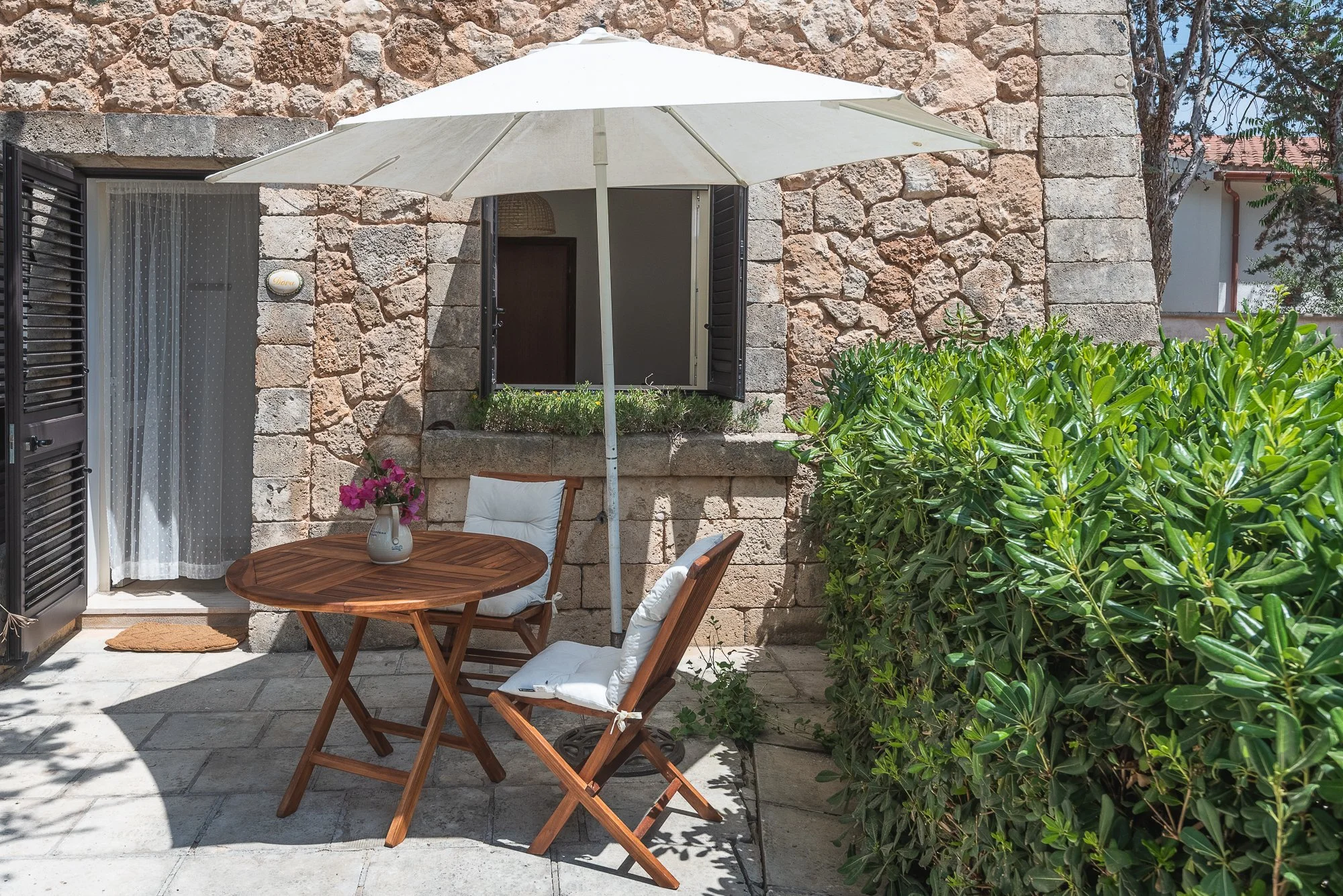 Ein Garten mit einem runden Holztisch, zwei Holzsessel mit weißen Kissen, einem weißen Sonnenschirm, einer Steinmauer und einer großen grünen Hecke.