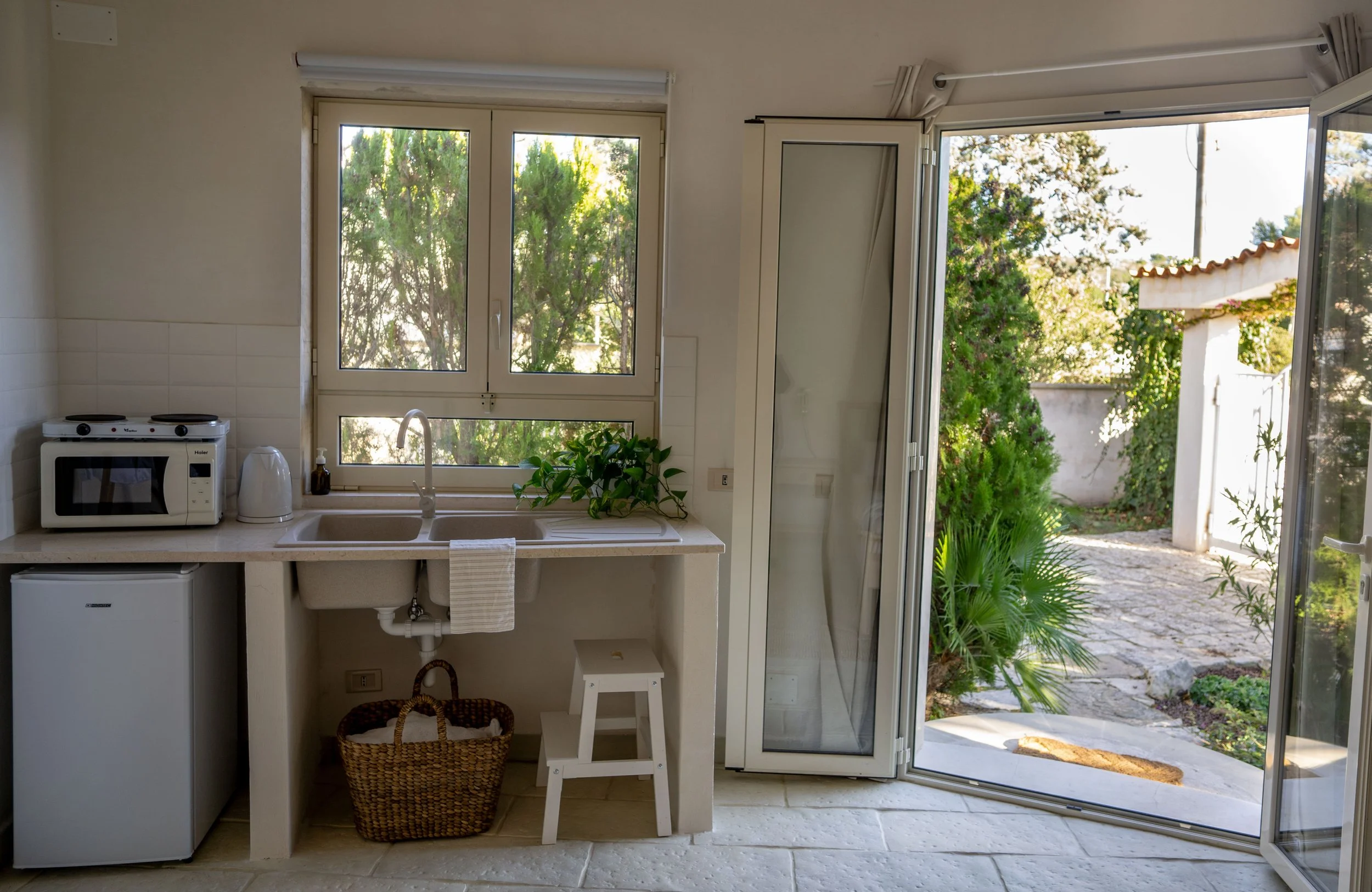 Küchenbereich mit Fenster, offener Tür nach draußen, kleine Mikrowelle, Wasserkocher, Waschbecken, grüne Pflanzen, Blick ins Freie mit Gartenpflanzen und Steine
