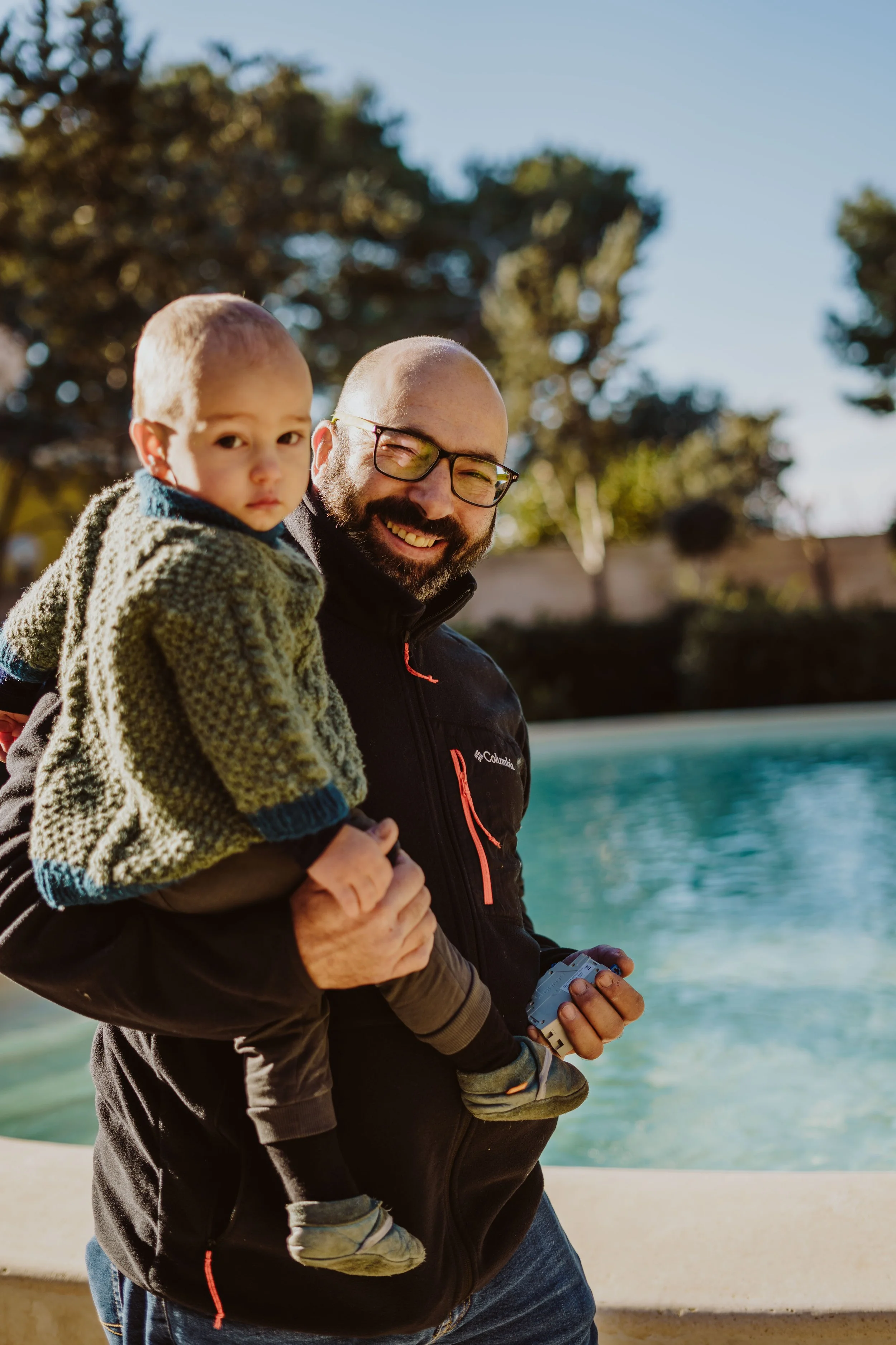 Ein Mann trägt einen kleinen Jungen auf dem Arm, vor einem Wasserbecken im Freien an einem sonnigen Tag.