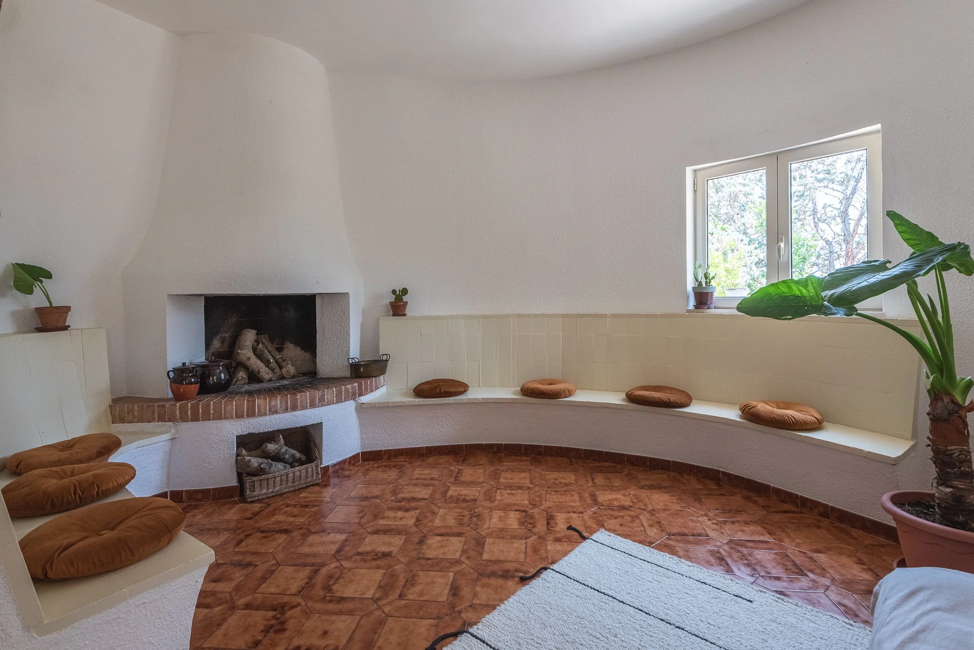 Gemütlicher Raum mit weißen Wänden, braunen Fliesen, Sitzecke mit braunen Kissen, Kamin, eine Fensterbank mit Pflanzen