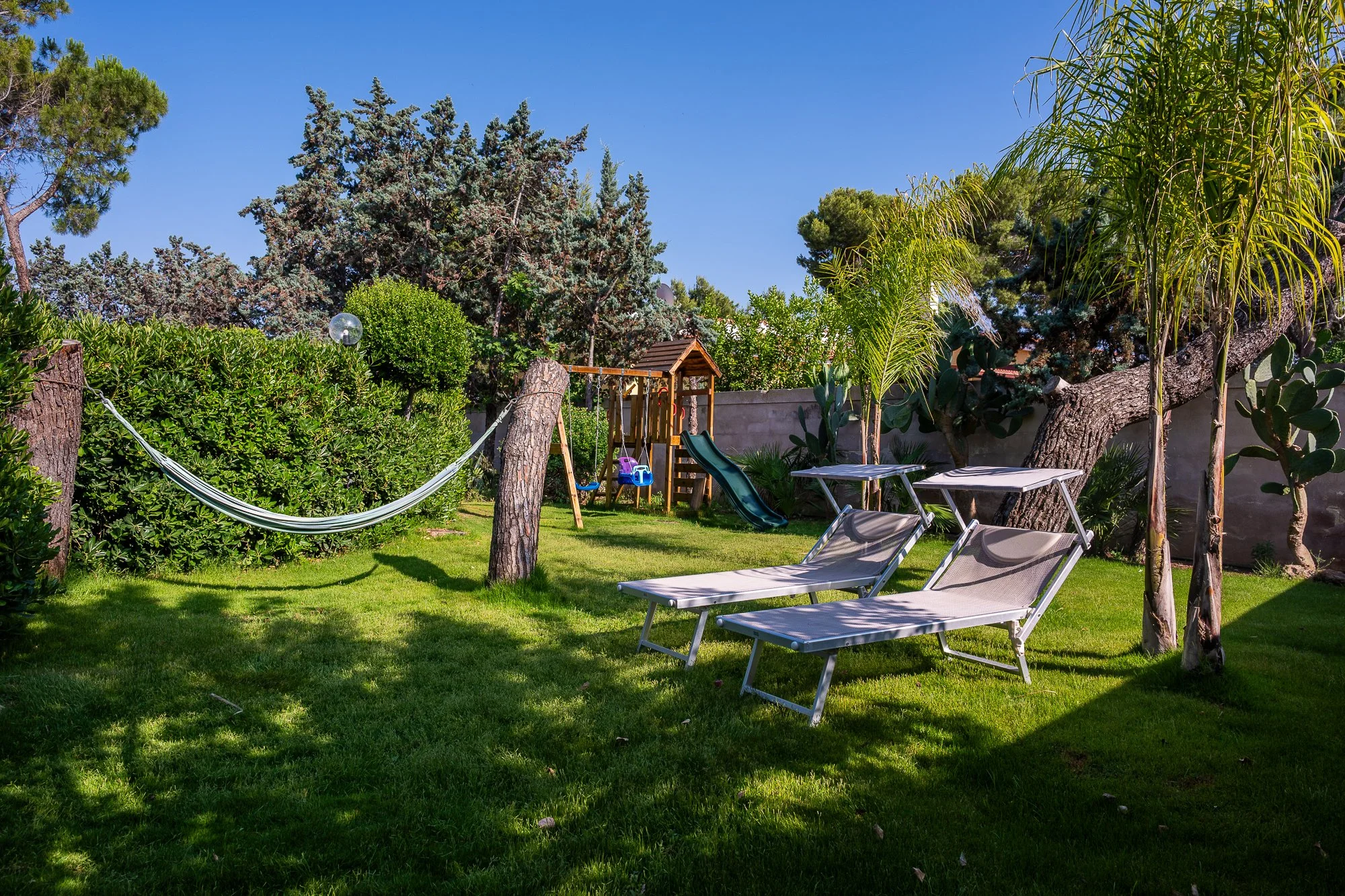 Garten mit Liegestühlen, Schaukel, Hängematte, Bäumen und verschiedenen Pflanzen unter blauem Himmel.