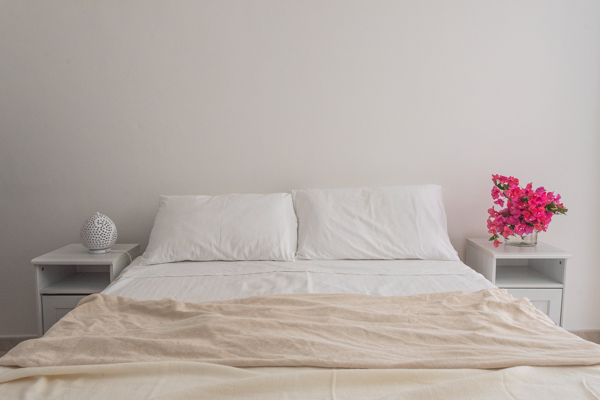 Bett mit weißen Kissen und Decke, zwei Nachttische mit Blumen und Lampe auf einem weißen Wandhintergrund.