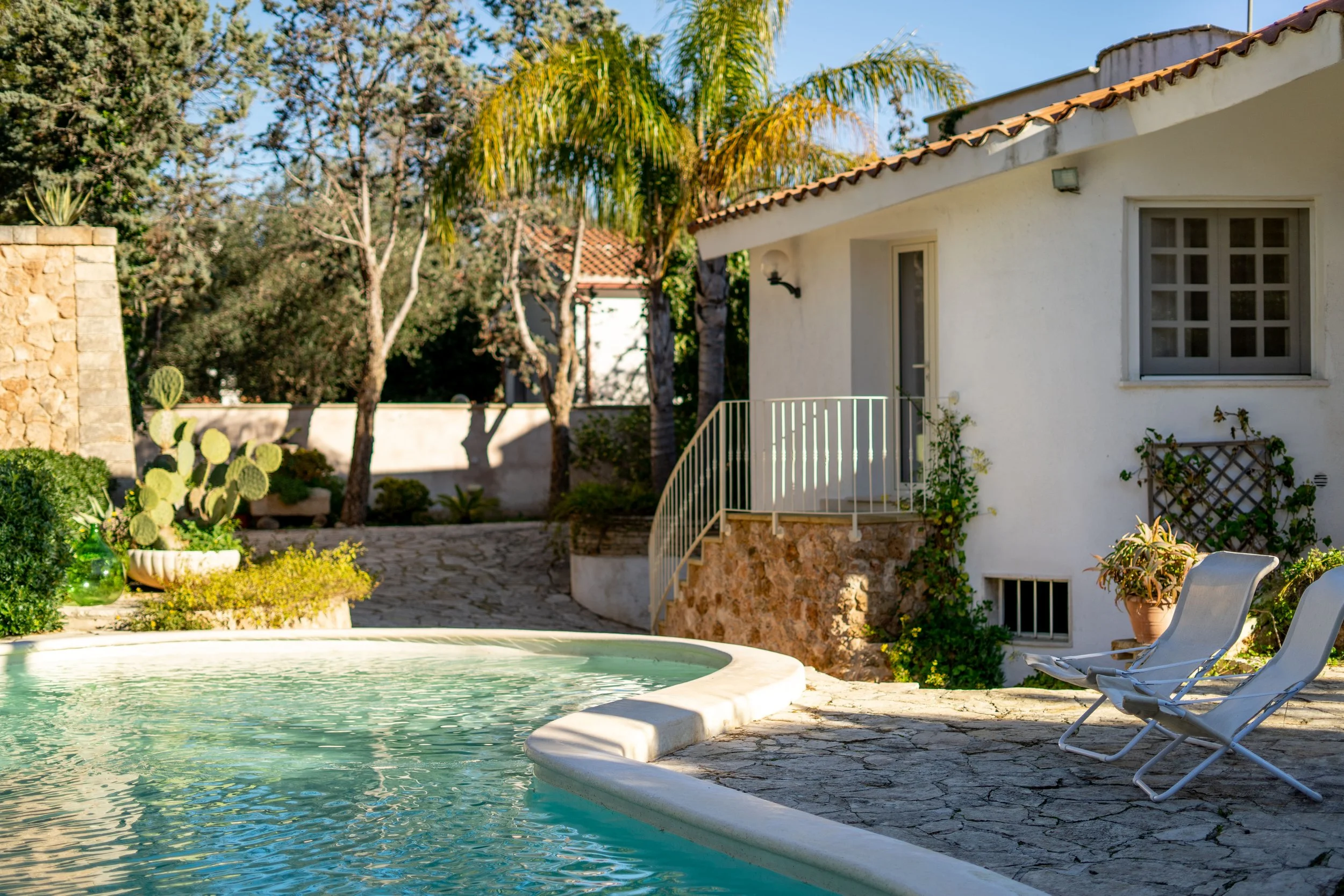 Ein Pool im Hintergrund eines kleinen weißen Hauses mit Terrasse, umgeben von Pflanzen und Bäumen, bei sonnigem Wetter.