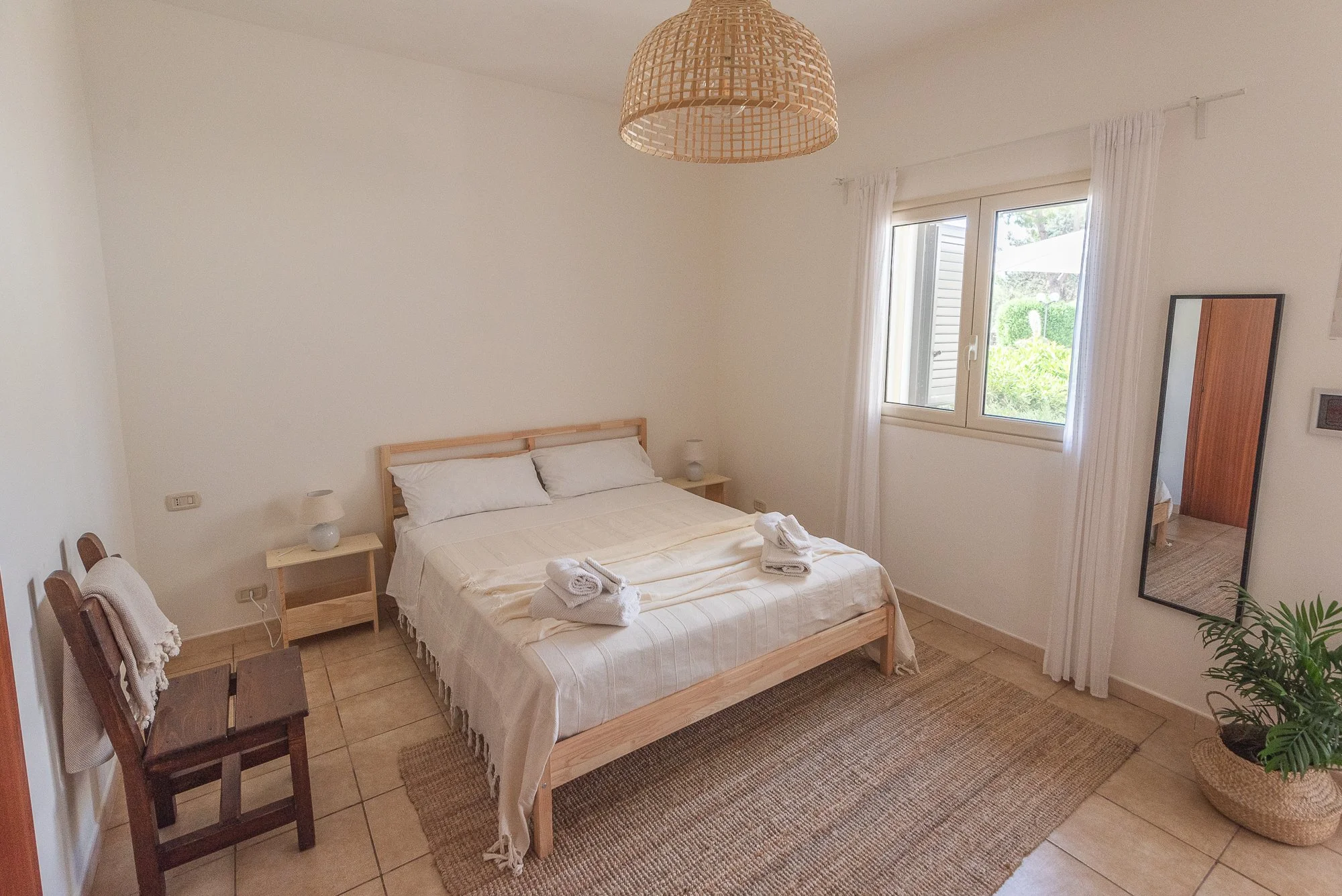 Ein Schlafzimmer mit Doppelbett, weißen Bettwäsche, zwei Nachtschränkchen mit Lampen, einem Spiegel, einem kleinen Stuhl mit Handtüchern und einem Teppich.