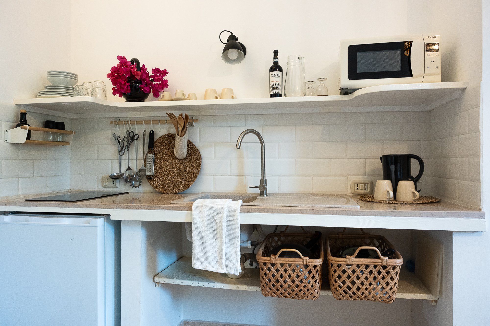 Küchenutensilien, ein Wasserkocher, Geschirr, und eine Mikrowelle auf einem weißen Küchenregal, darunter Körbe für Lebensmittel oder Küchenutensilien.