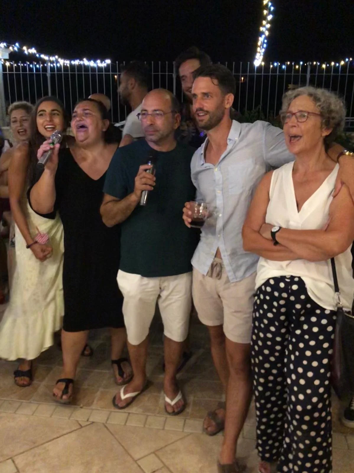 Gruppe von Menschen bei einer Party nachts, einige singen, andere trinken, im Hintergrund Lichterketten.