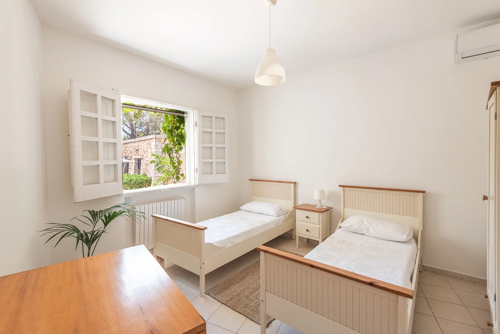 Schlafzimmer mit zwei Einzelbetten, einem Nachttisch, einem Lampen, offenen Fenster mit Blick auf Bäume, Pflanzen und Gebäude, weiß gestrichene Wände.