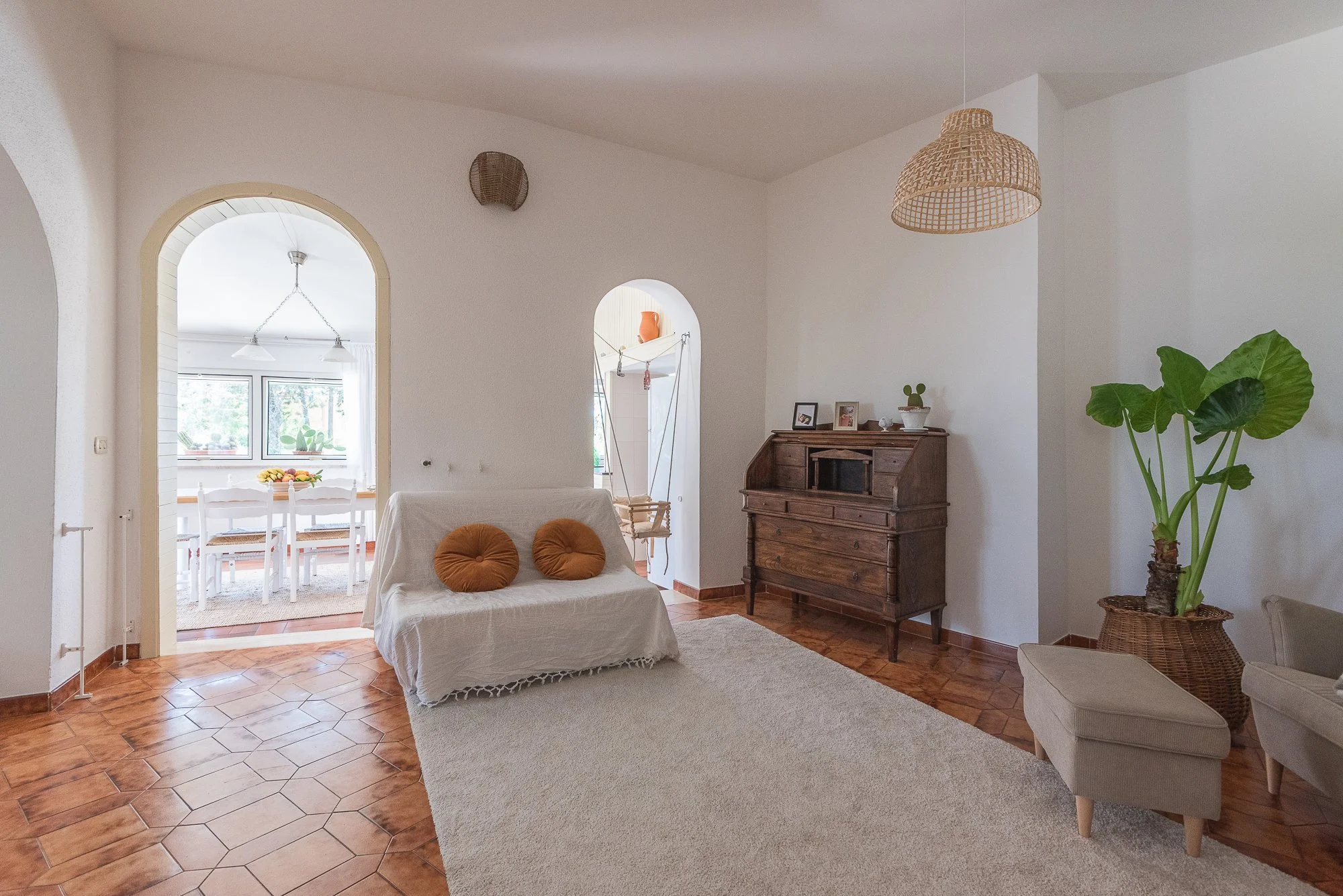 Ein helles Wohnzimmer mit braunem Holzfußboden, weißer Wandfarbe, einem weißen Sofa mit zwei braunen Kissen, einem antiken Holzsekretär, einer großen Pflanze in einem Korb und einem beige-hellgrauen Sitzhocker