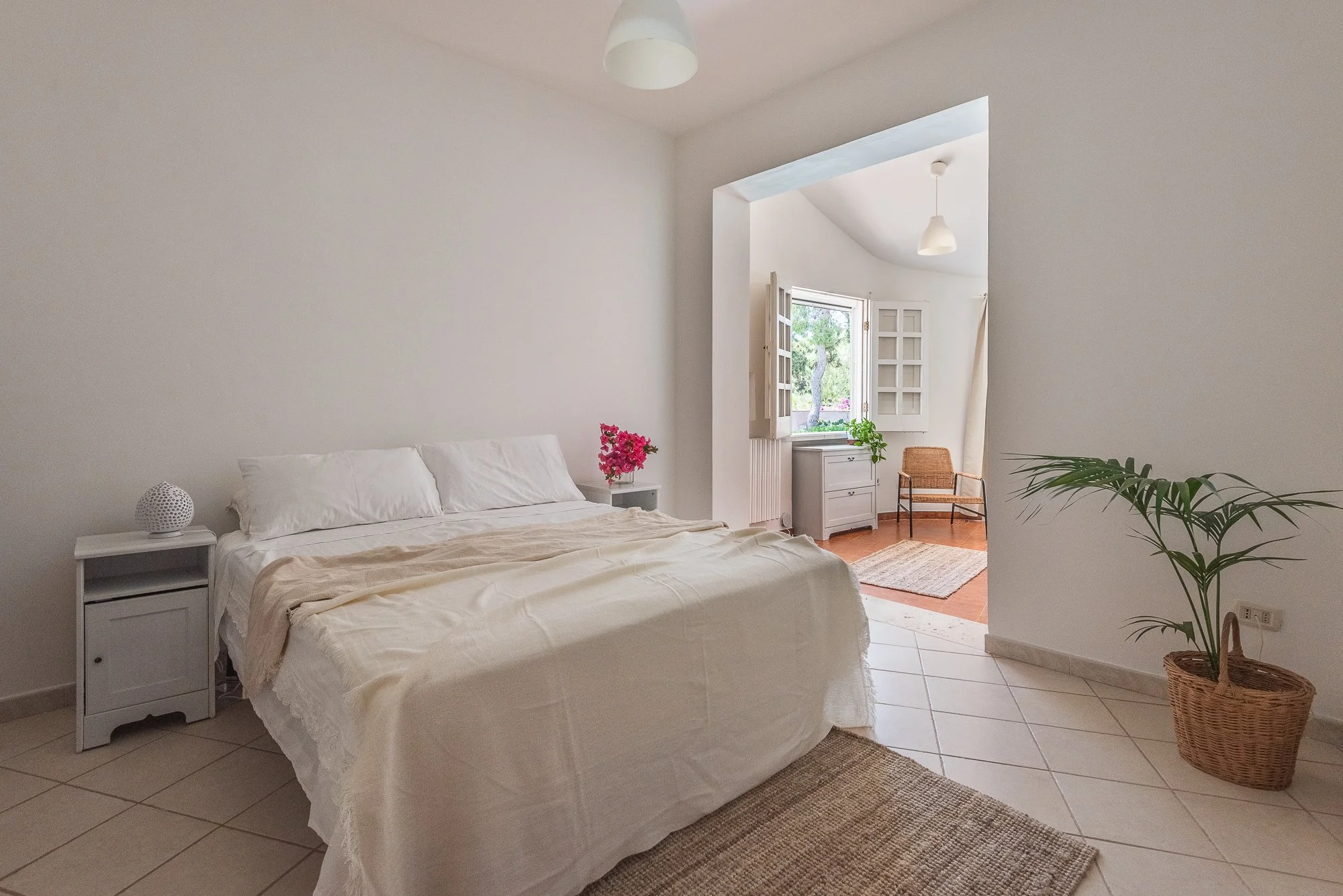 Schlafzimmer mit weißen Wänden, einem Bett mit weißen Bettwäsche und einem Nachttisch auf der linken Seite. Im Hintergrund ist ein Fenster mit offenen Fenstertüren, die auf einen Balkon oder Außenbereich führen, mit einem Stuhl und einem kleinen Tisc