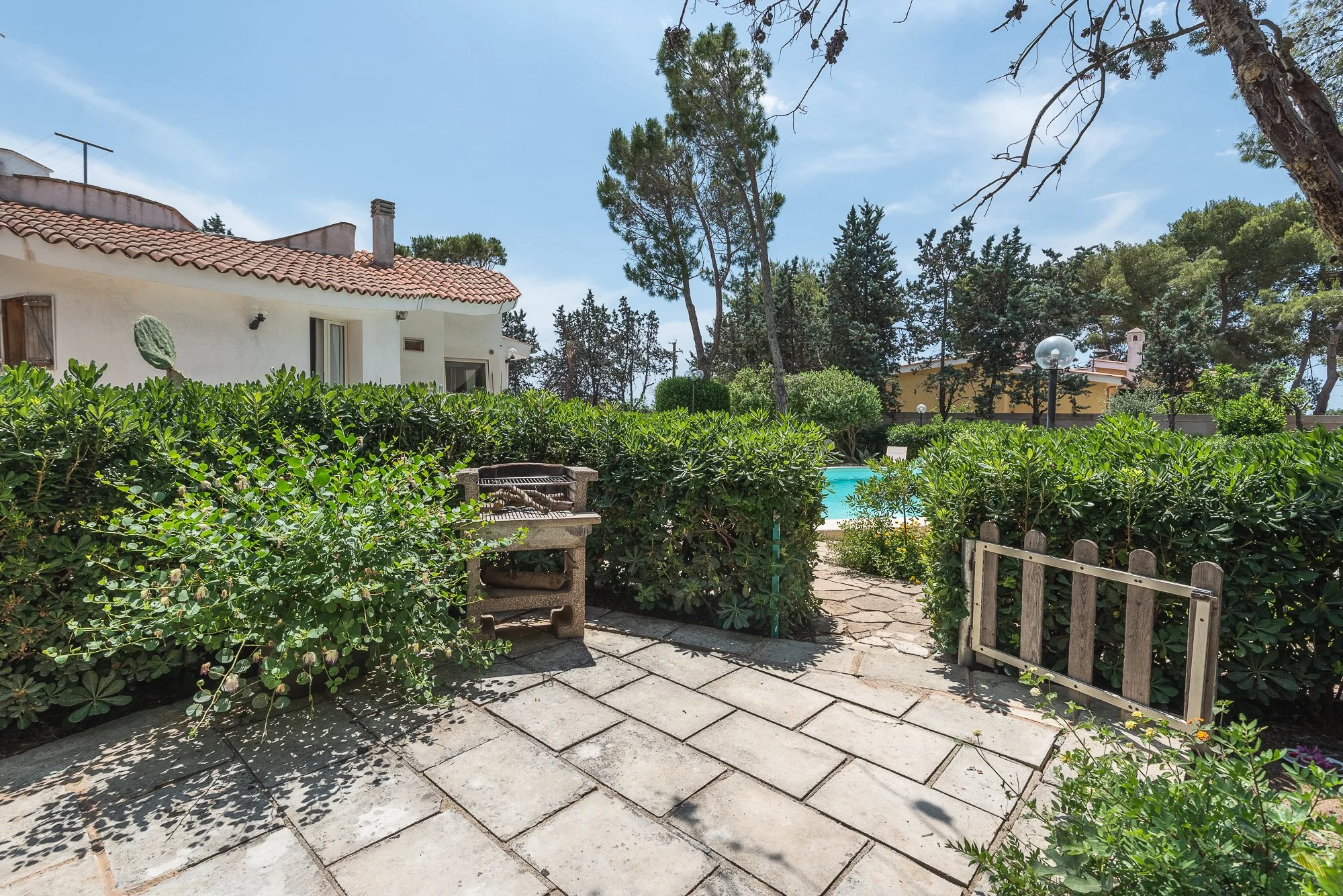Ein Garten mit gepflastertem Weg, grüne Büsche, ein Grill, ein Pool im Hintergrund, umgeben von Bäumen und einem Haus mit rotem Ziegeldach.