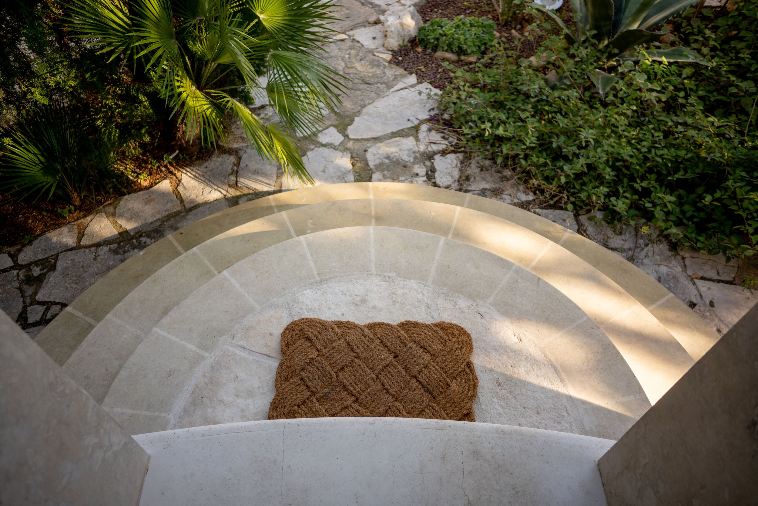 Blick nach unten auf eine steinerne, semi-kreisförmige Stufe mit einem braunen, geflochtenen Fußmatte in der Mitte, umgeben von grünen Pflanzen und einem gepflasterten Weg.