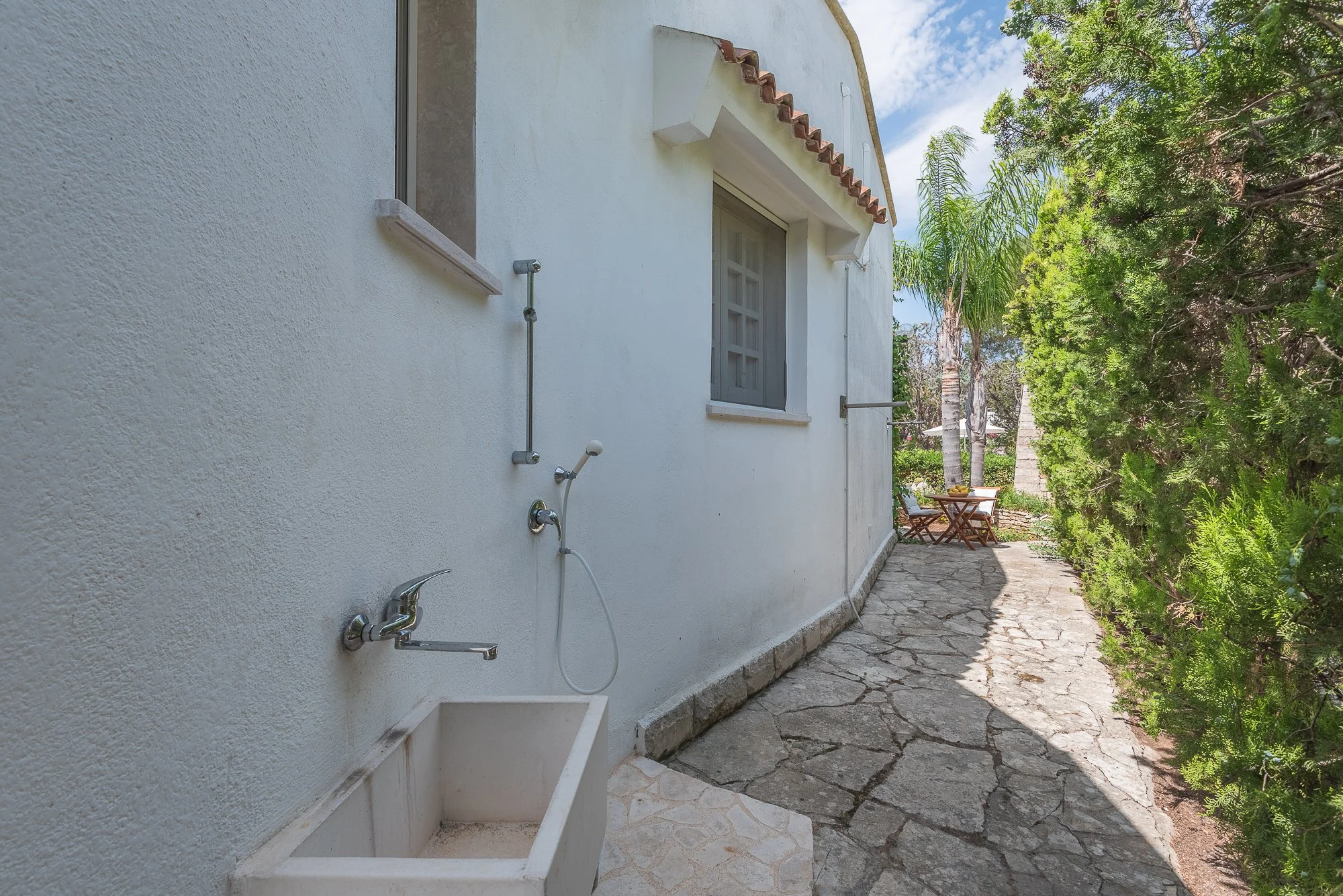 Ein weißes Haus mit einem kleinen Außenwaschbecken und einem Fenster, umgeben von grünen Bäumen und einem Steinweg, im Hintergrund ein Sitzbereich mit einem Tisch und Stühlen.